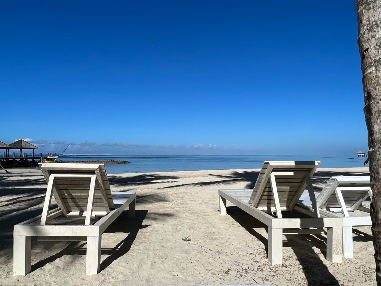 View past lounge chairs and white sand beach to a flat lagoon. 