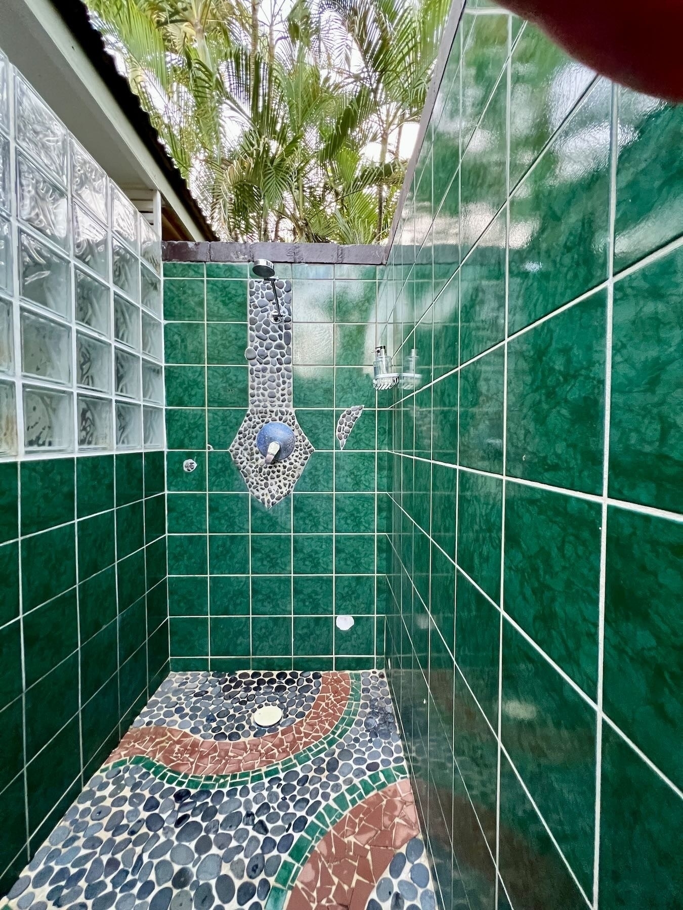 Tiled shower with walls but no roof. Beatiful patters of stones make up the floor. Palm trees are visible. 