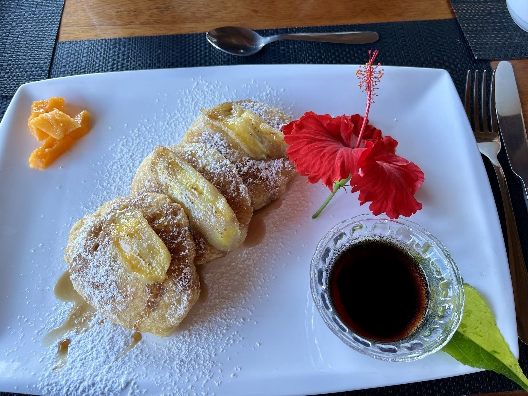 Pancakes with banana on top and a bowl of syrup. A red flower decorates the plate. 