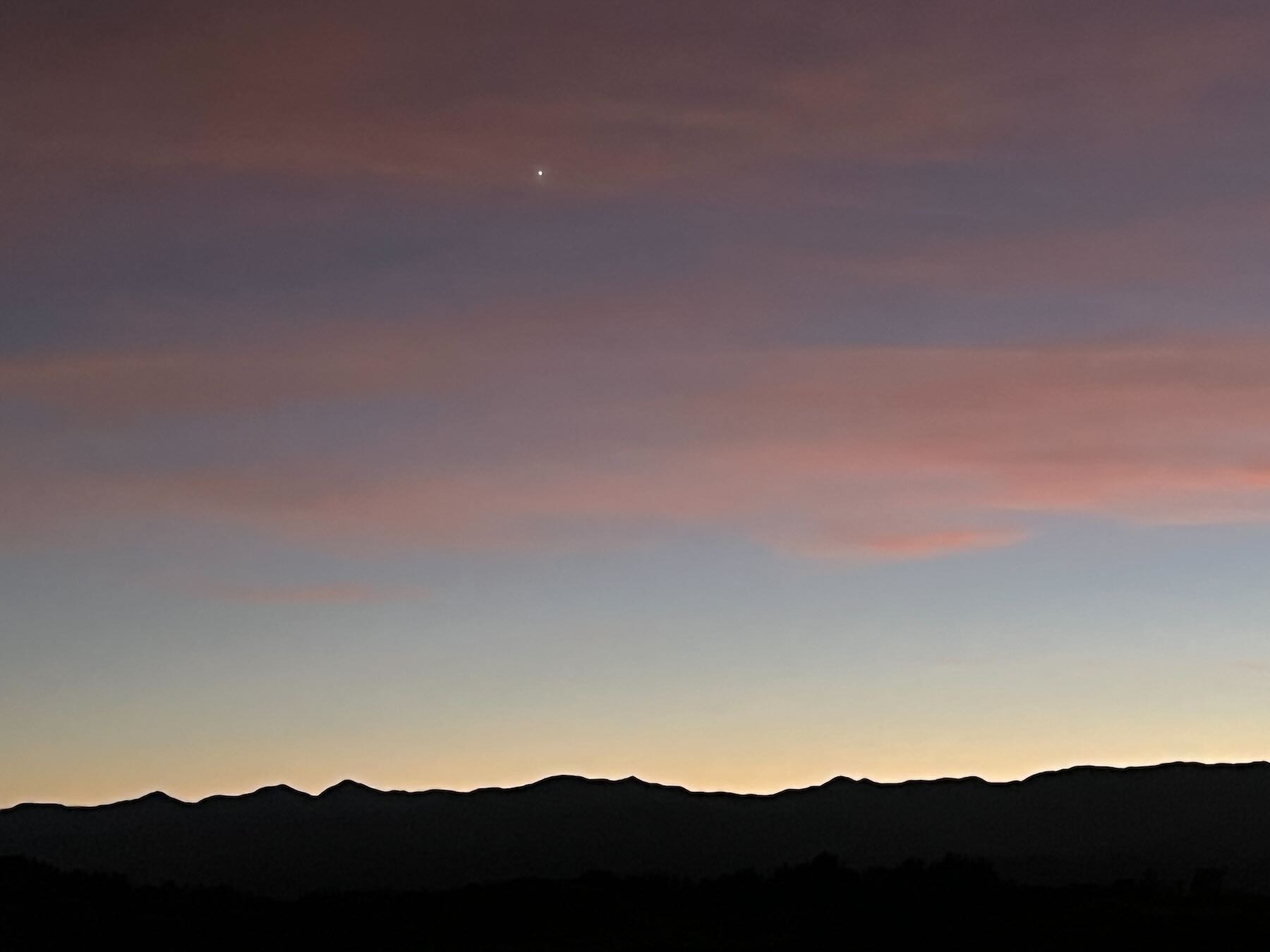 Venus high above shadowy mountains, hazy pink cloud streaks the sky. 