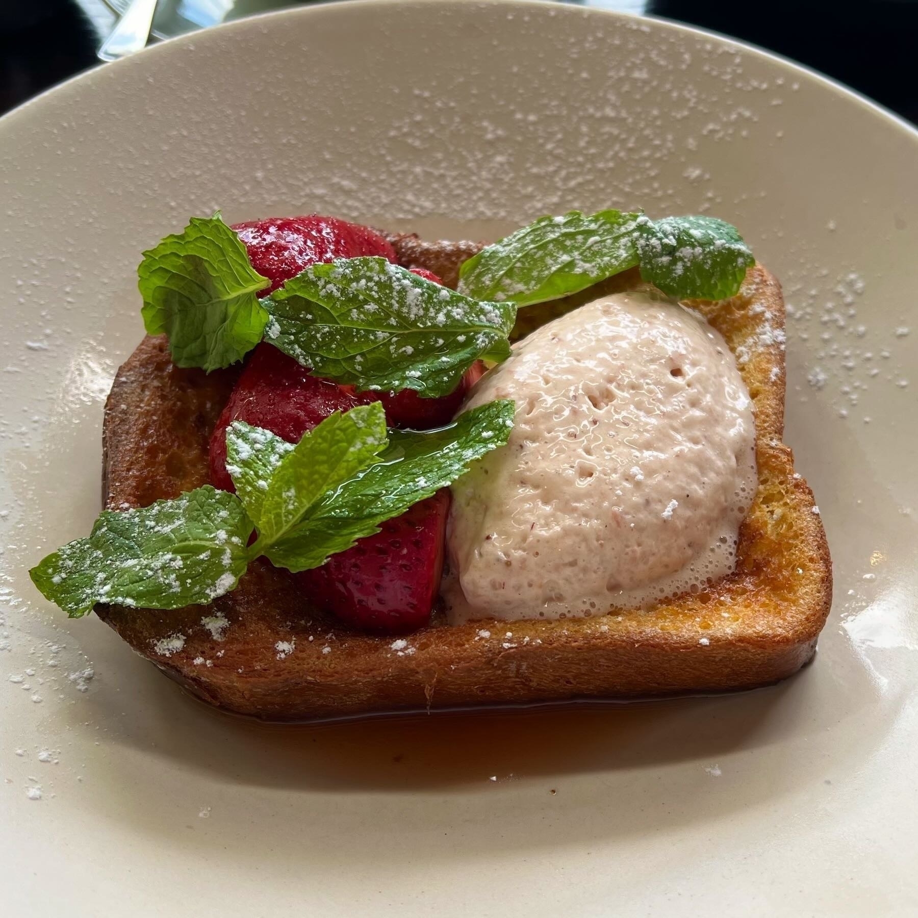 White plate with a slice of brioche, strawberries, and a cream like substance, topped with green leaves. 