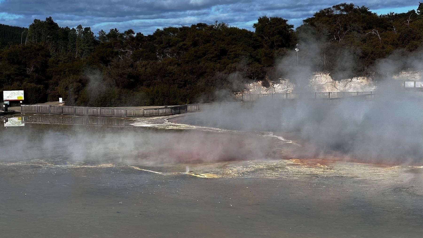 Pool with steam, white and yellow colours, with red showing through. There are paths and trees in the background. 