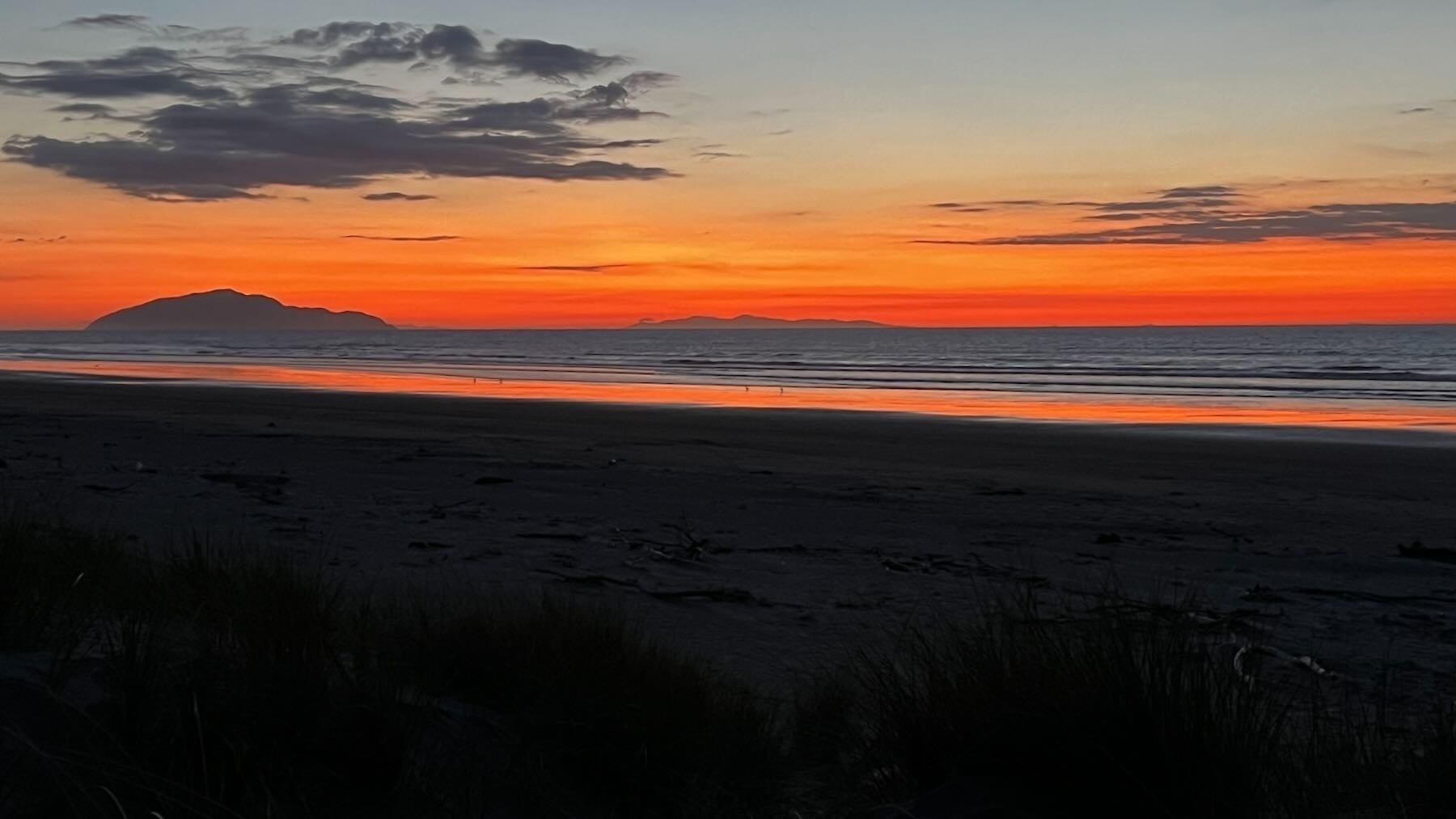 Dark below, then pink wet sand, then an island beside a startling bluish sea, with scarlet above, then cloud, then blue sky.