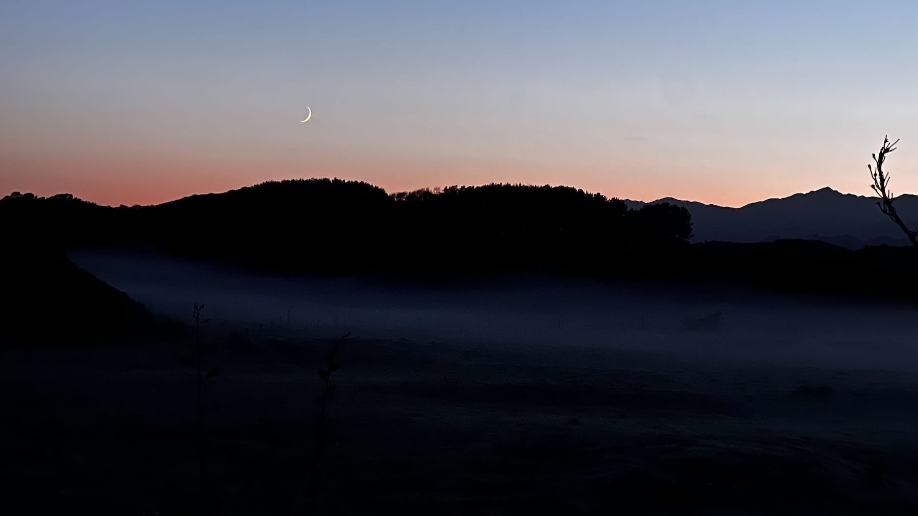 Crescent moon above dark hills, with a bit of gound fog in the foreground.