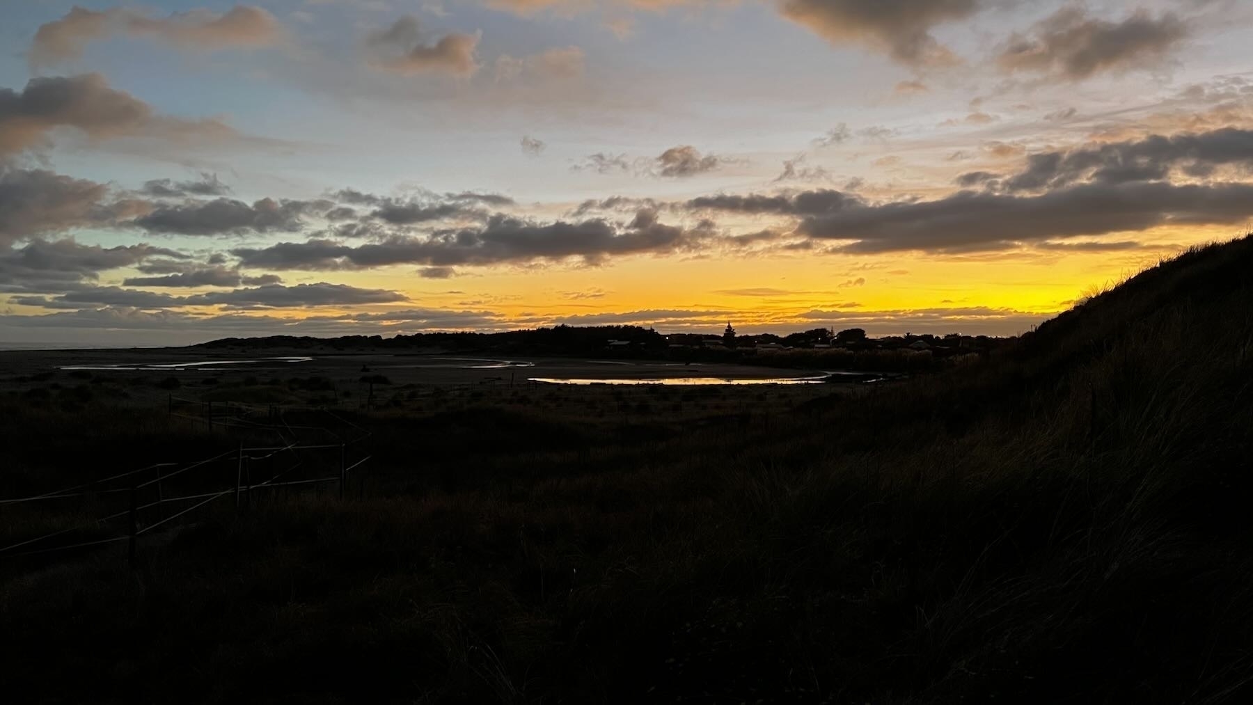 A small bright spot of water in an otherwise datk landscape with pale blue and orange sky between clouds. 