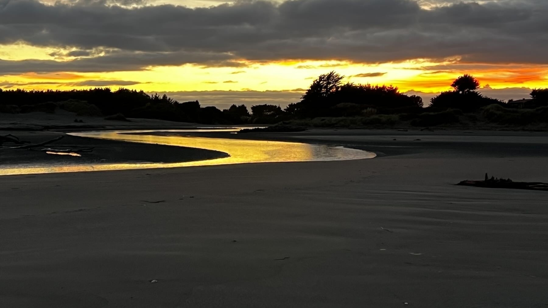 A yellow ribbon of river winds through a darker beach landscape as the sun starts to light a partly cloudy sky with orange. 