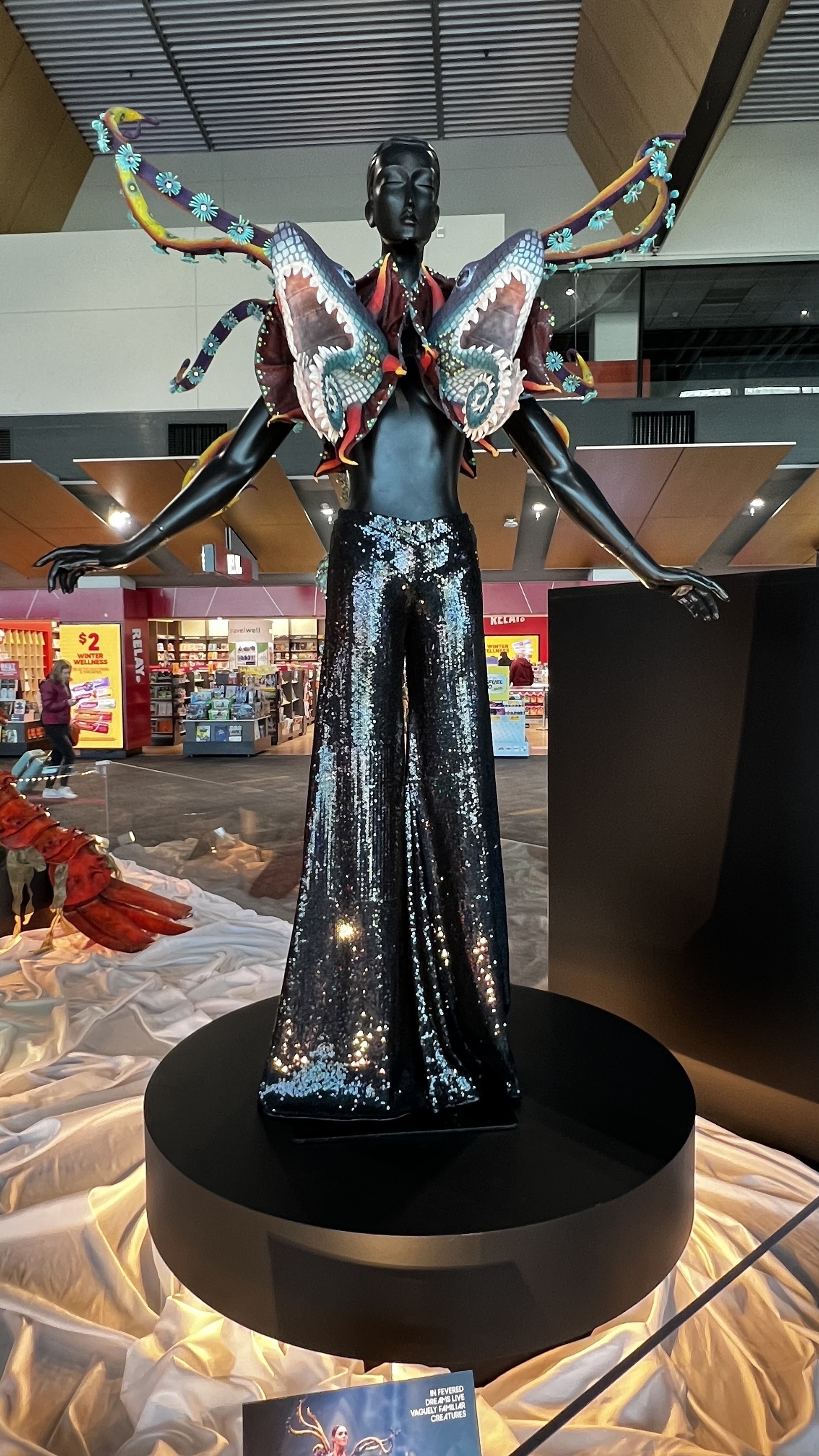 A mannequin dressed in a shimmering outfit with an elaborate butterfly-themed wing accessory is displayed on a pedestal, set against an open indoor background with visible shops and decor.