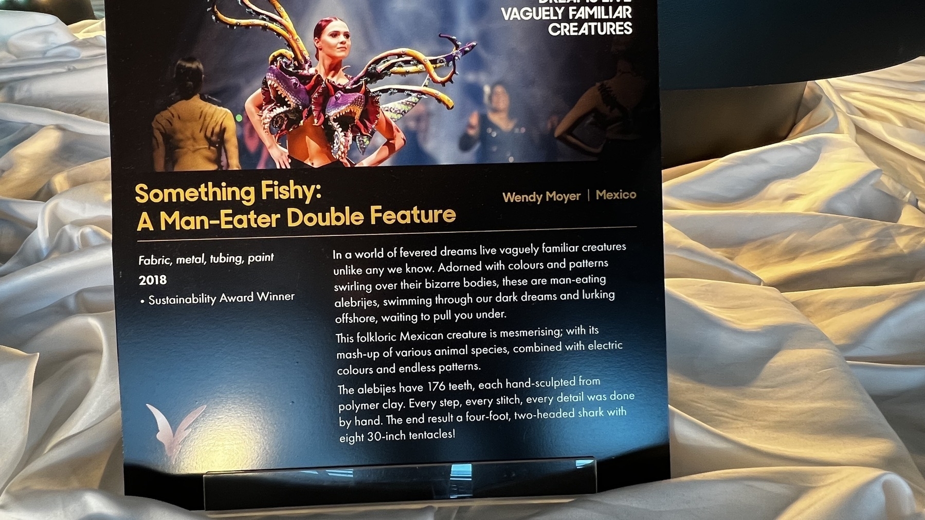 A display features a sign about an award-winning fabric and metal art exhibit titled "Something Fishy: A Man-Eater Double Feature" by Wendy Moyer from Mexico, set against a shimmering backdrop.