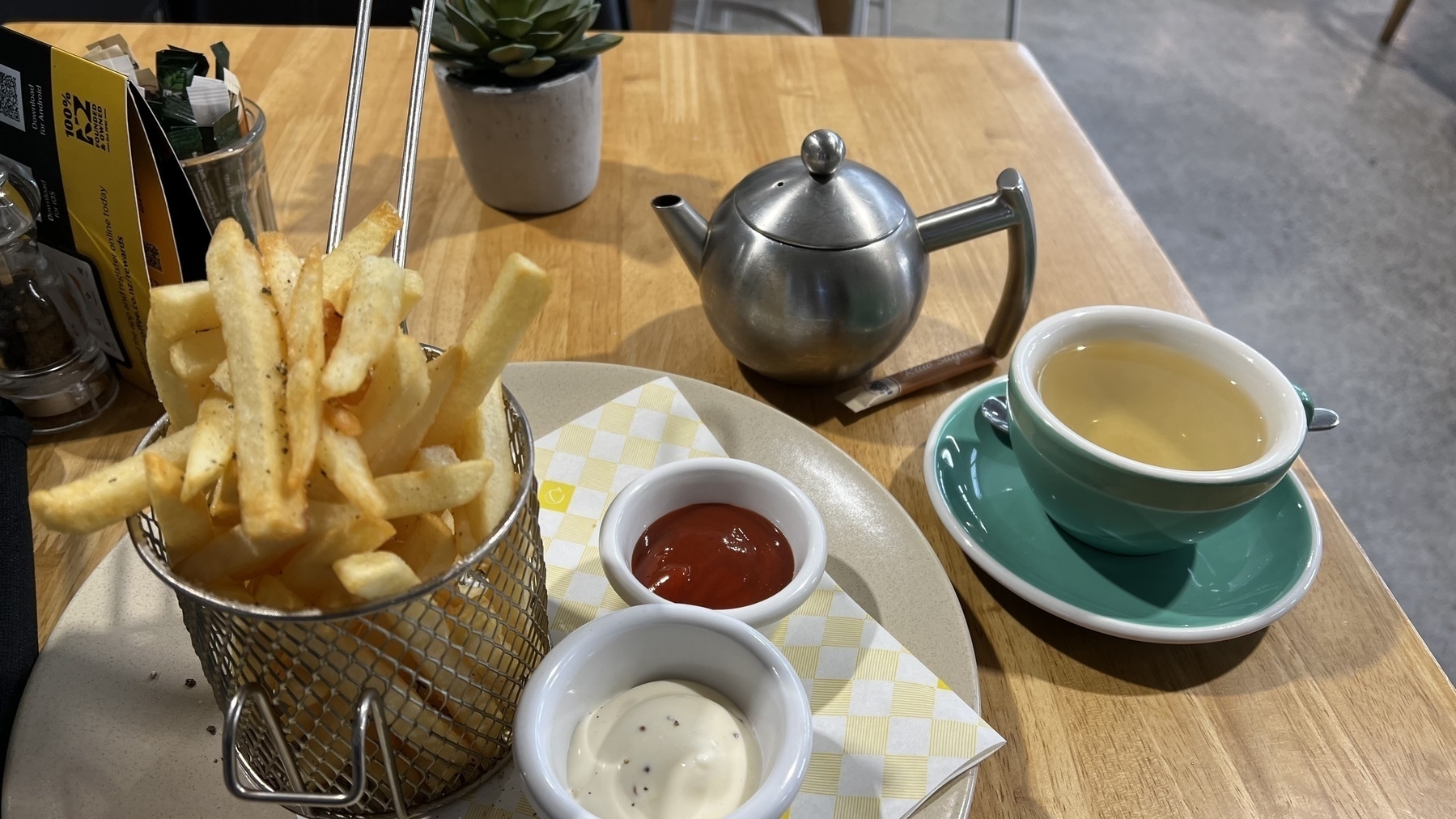 A serving of fries with ketchup and mayonnaise, a small tea pot, a cup of tea, and a potted plant are arranged on a wooden table.