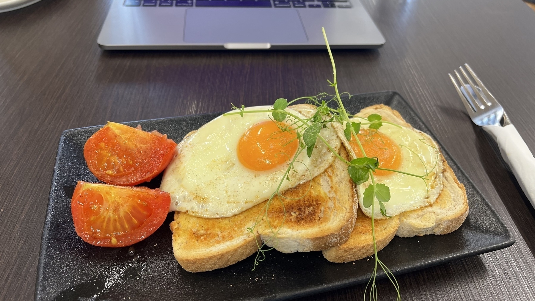 A plate of toast topped with sunny-side-up eggs and garnished with microgreens is served alongside sliced tomatoes, with a laptop in the background.