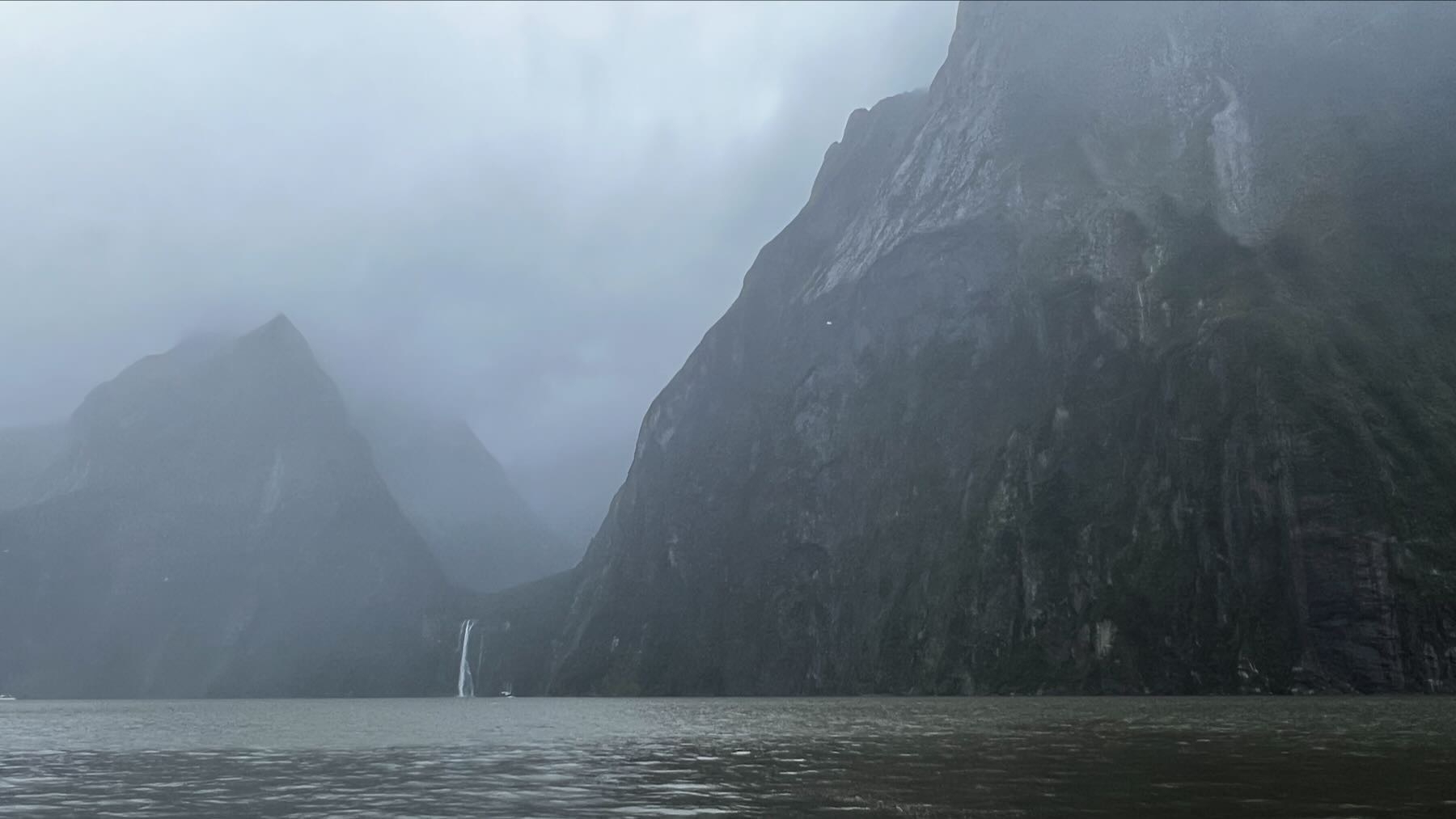 Huge misty bare mountains and a small waterfall, with a minute tourist boat at the base.