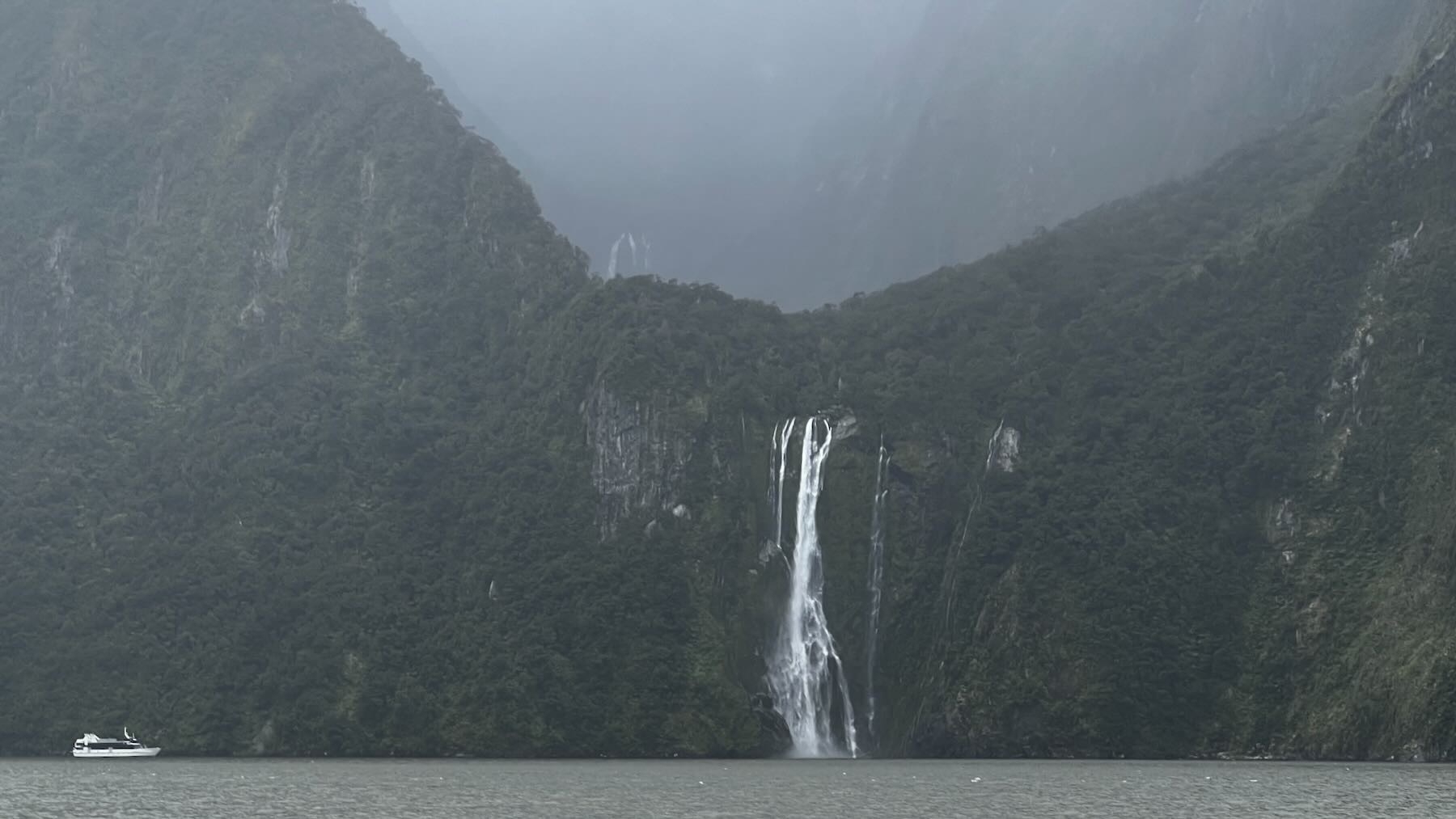 Closer view of a tall waterfall from bushclad misty mountains with a small tourist boat nearby.