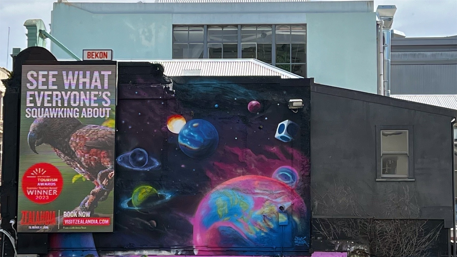 Mural shows colourful planets against a background of black. On the left an electronic billboard advertises Zealandia.