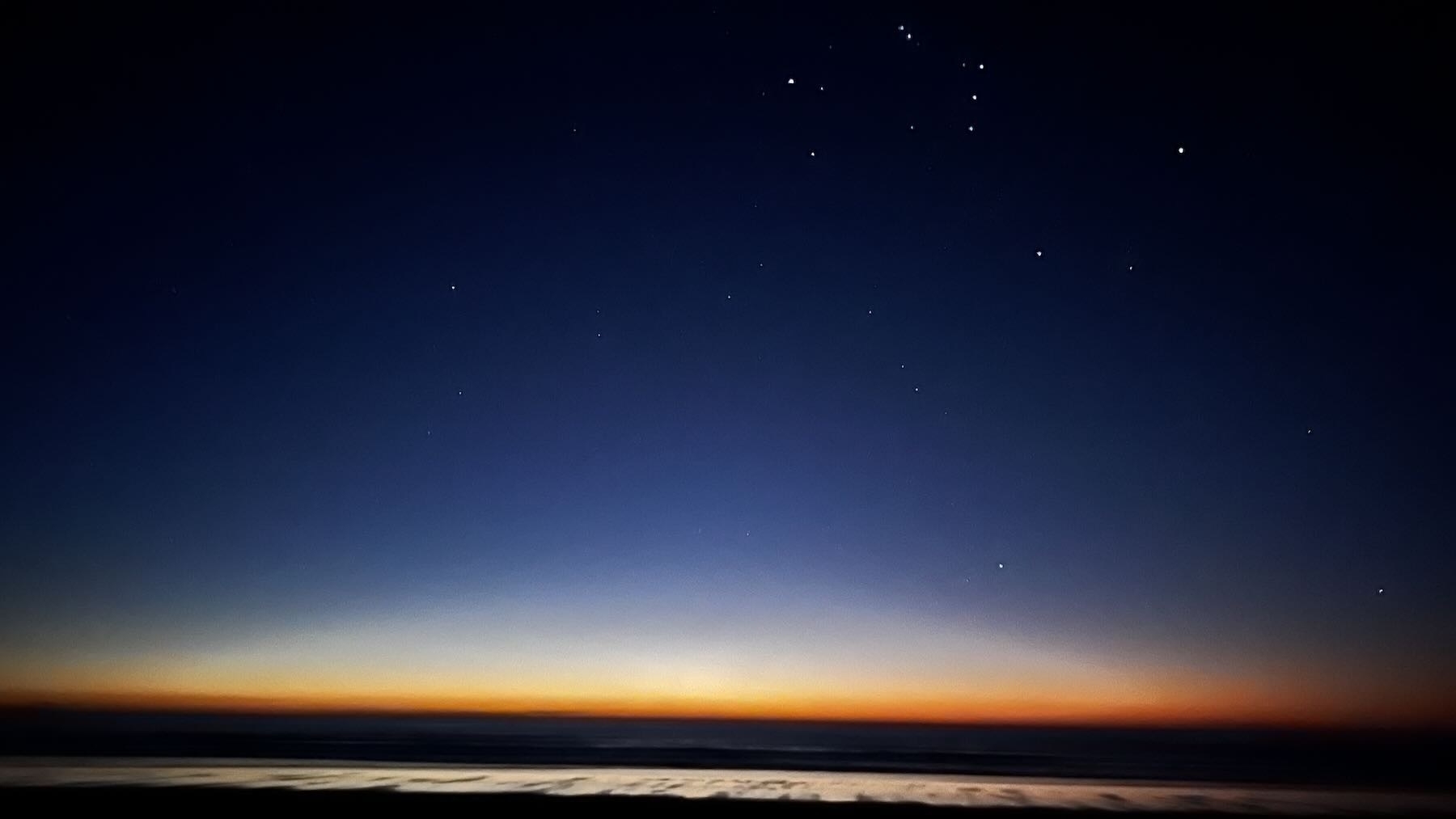 In the foreground a bright patch of damp sand, then a line of dar sea and a bright band of sky where the sun had recently set, with darker blue above in which the constellation of Orion can be made out.