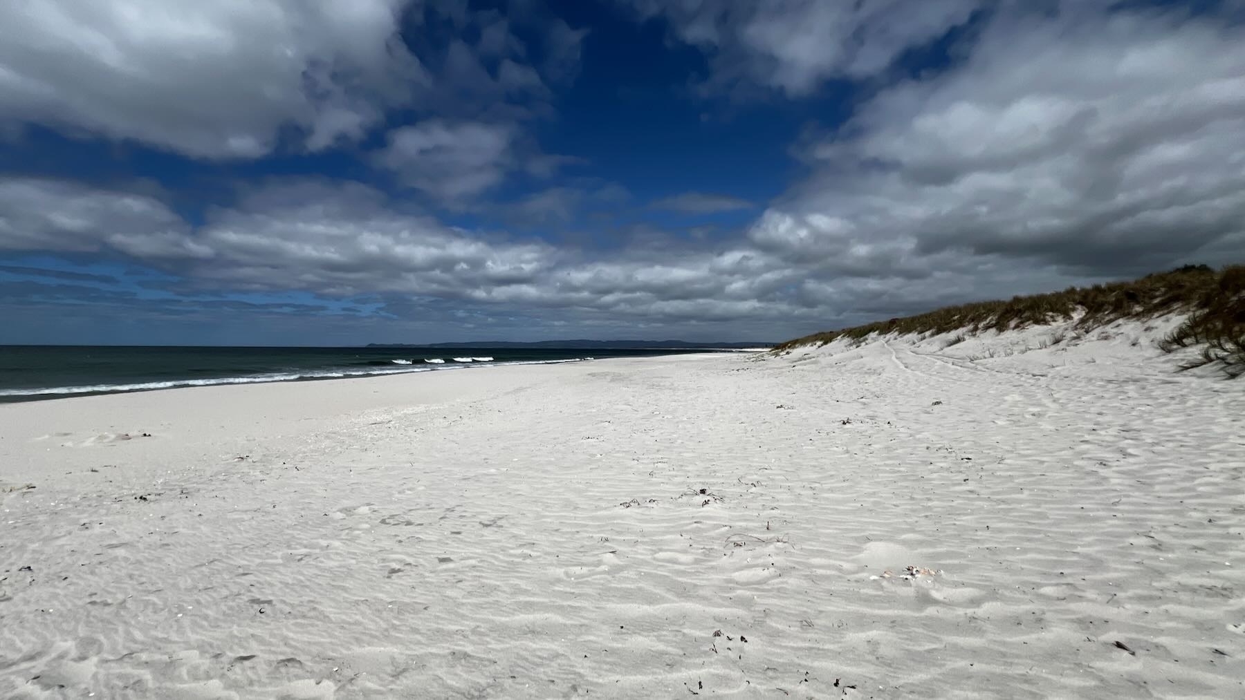 A white sand beach beneath a blue sky with scattered clouds. The sea is dark green. 