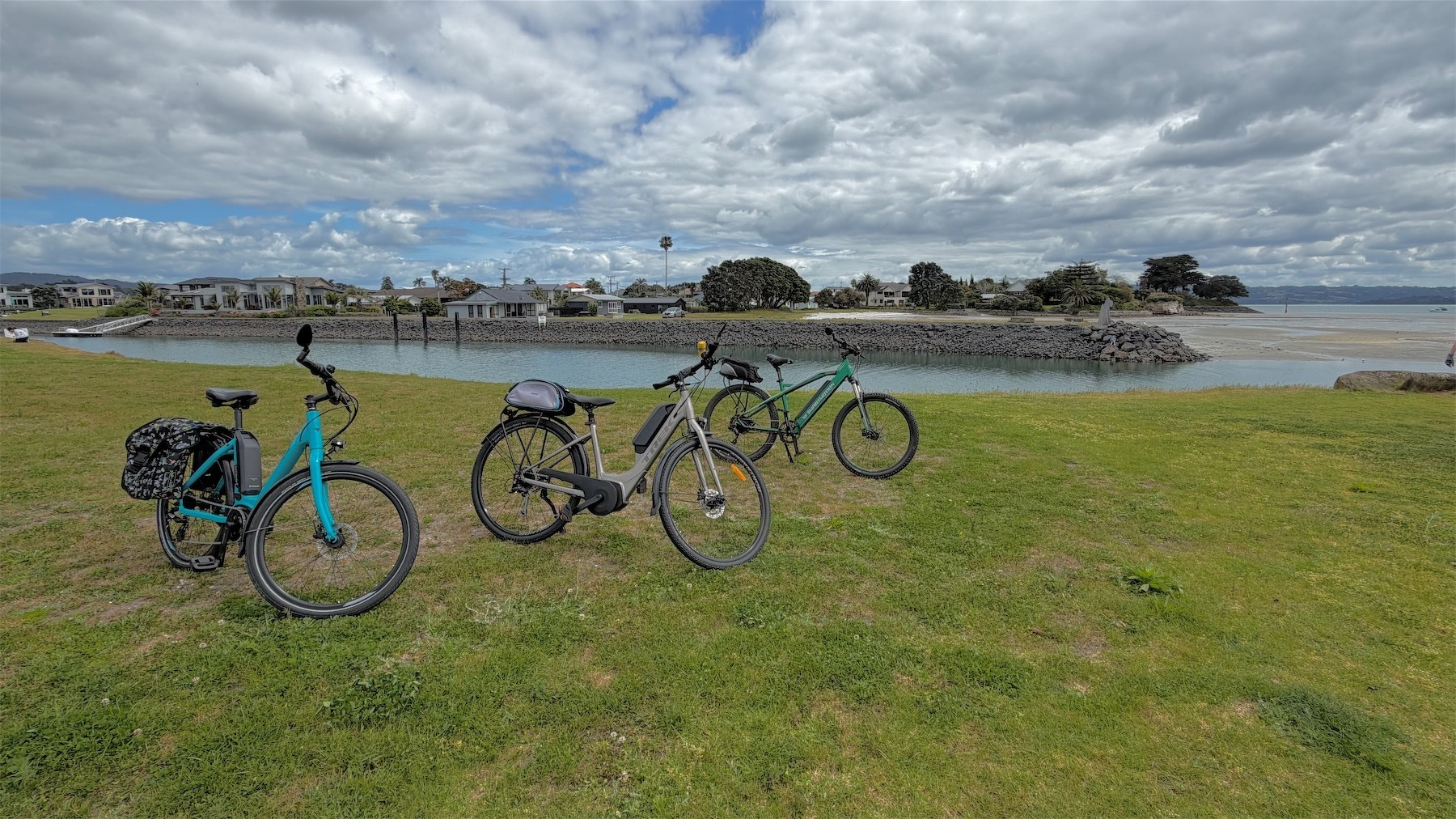 3 bikes on their stands on a grassy area. In the background a channel of water opening into a bay.