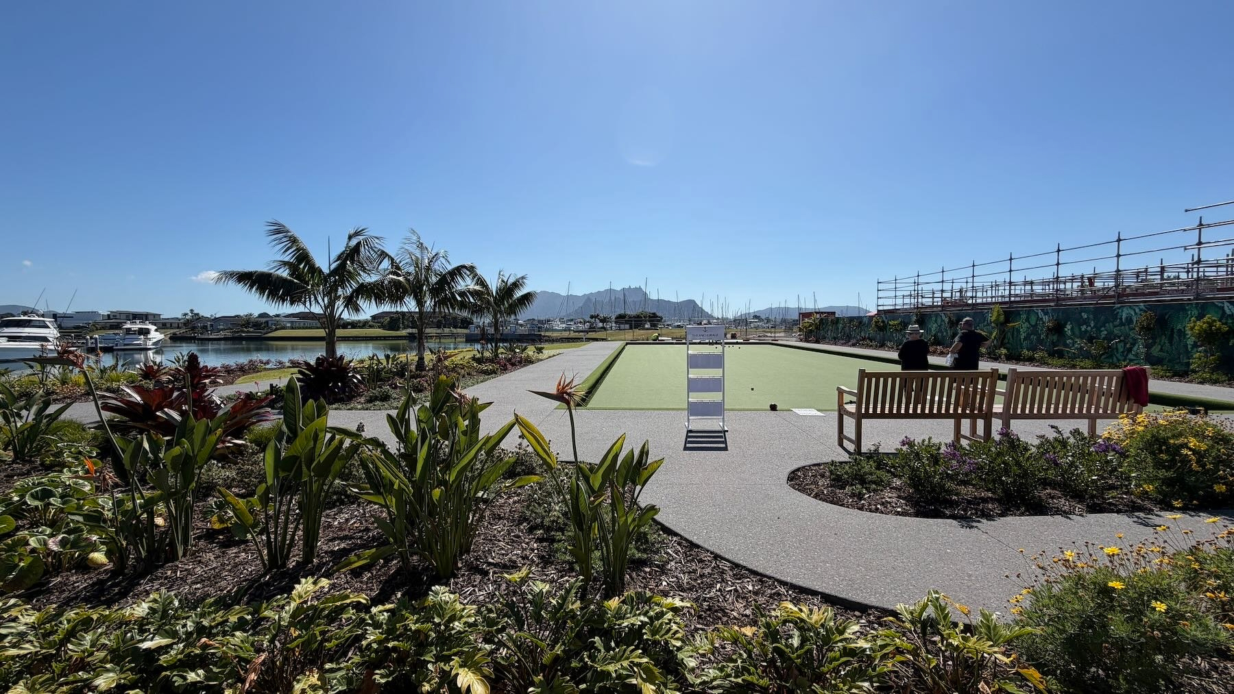 View across green gardens and a lawn bowls area to a marina with hills behind and a blue sky.