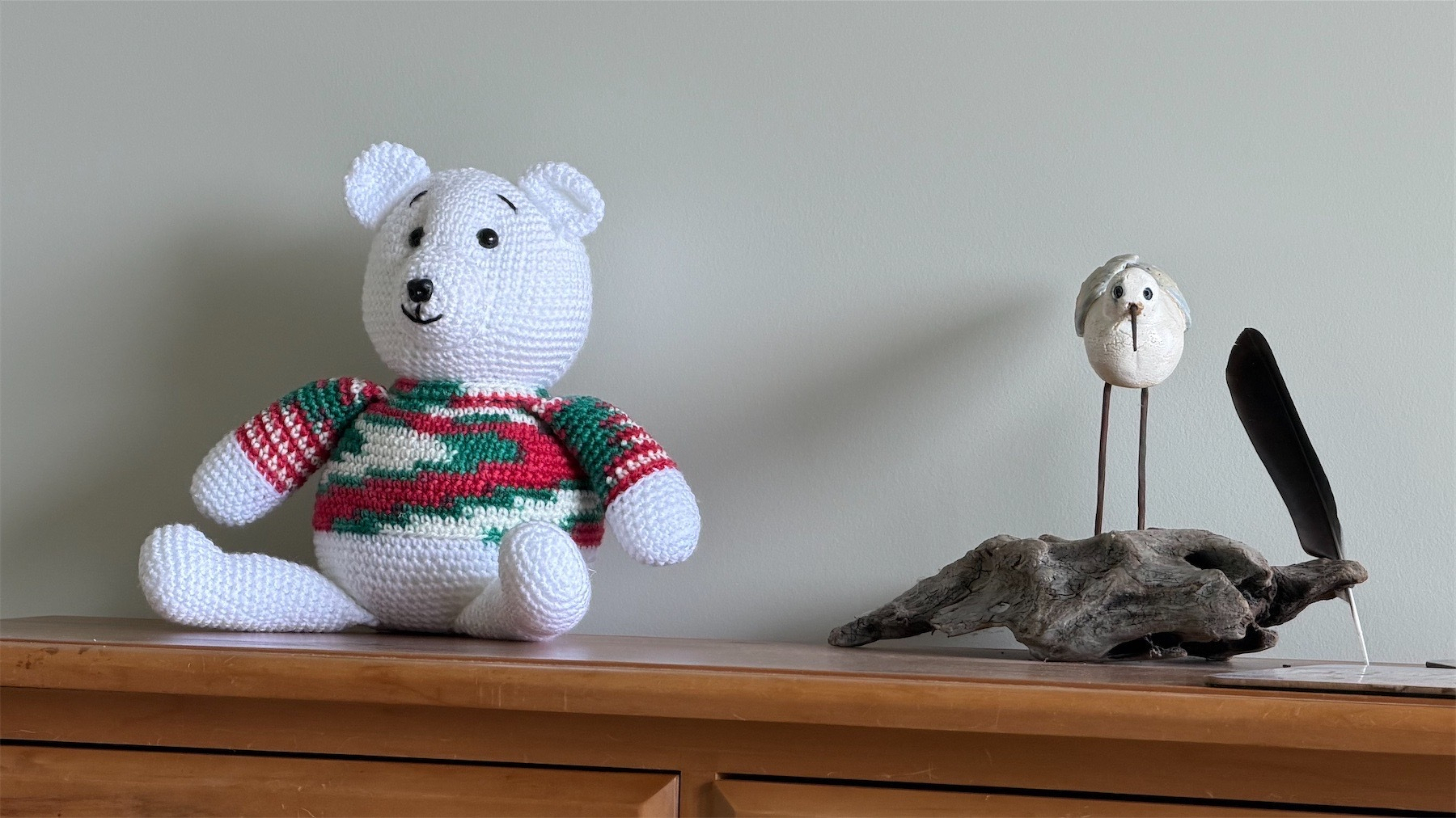 White knitted Bear in a Christmas jersey and bird sculpture atop a piece of wooden furniture.