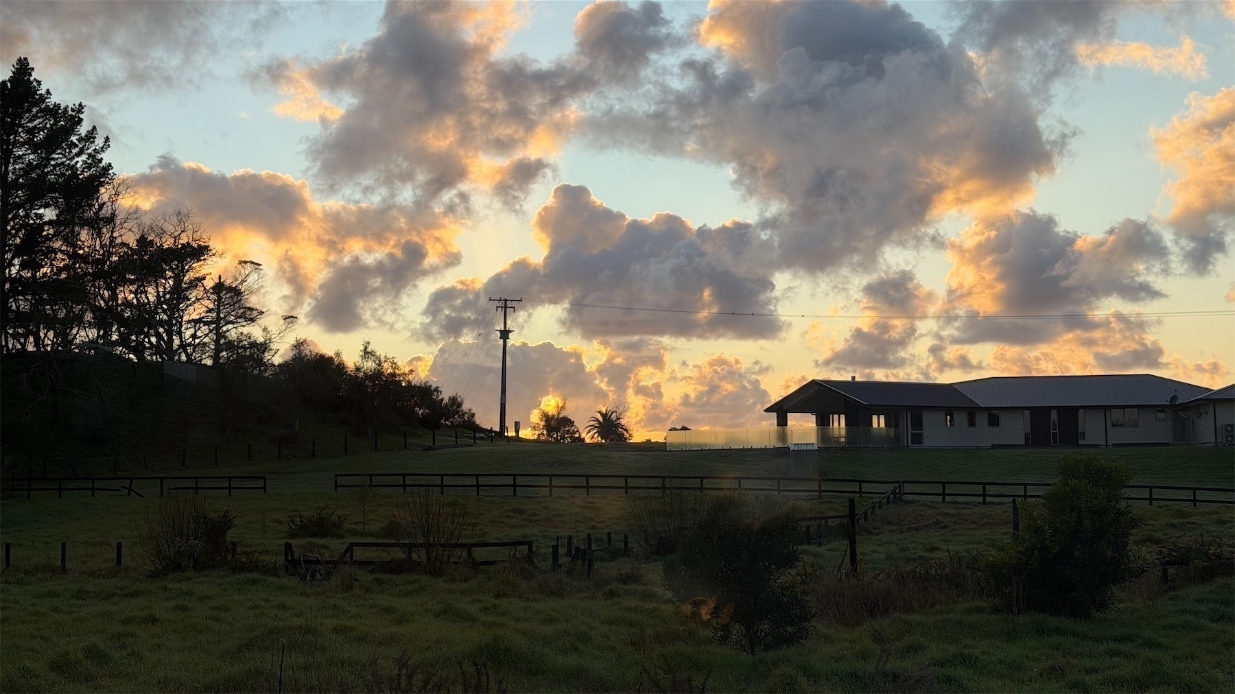 Dark grass and a house, with clouds behind, lit by a hidden rising sun.