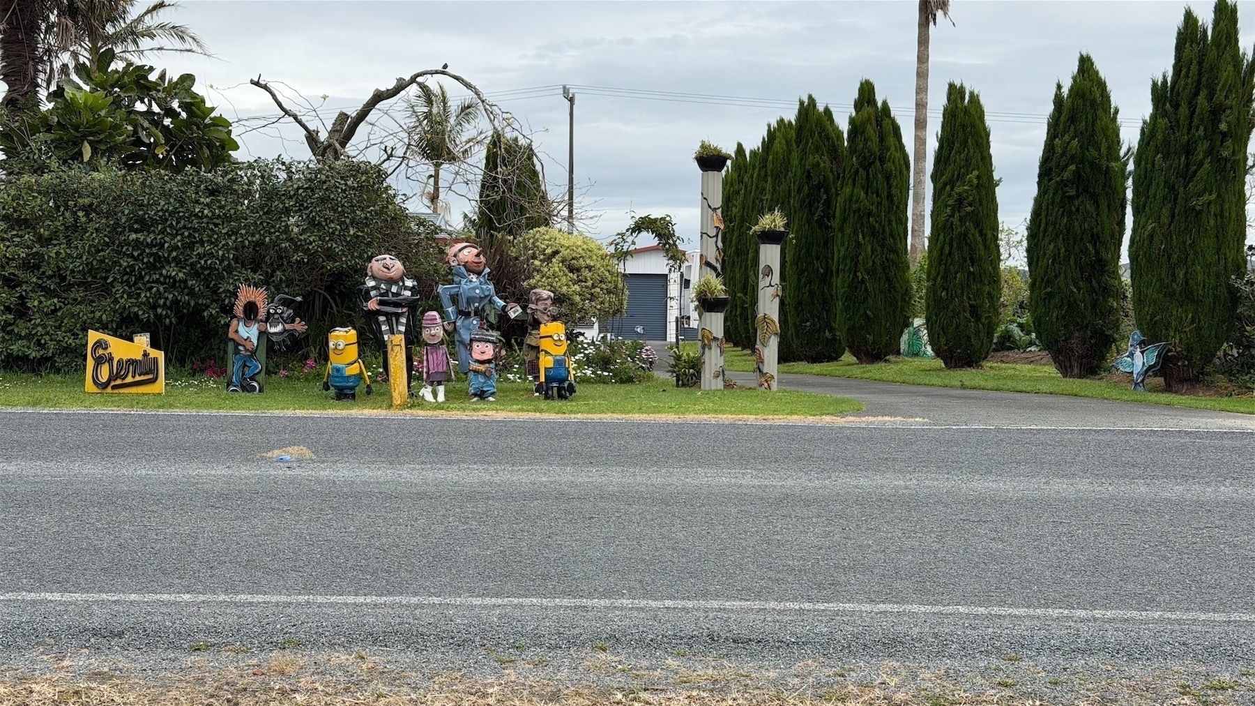 Several figures, including Minions, stand or sit before a hedge on the side of the road. 