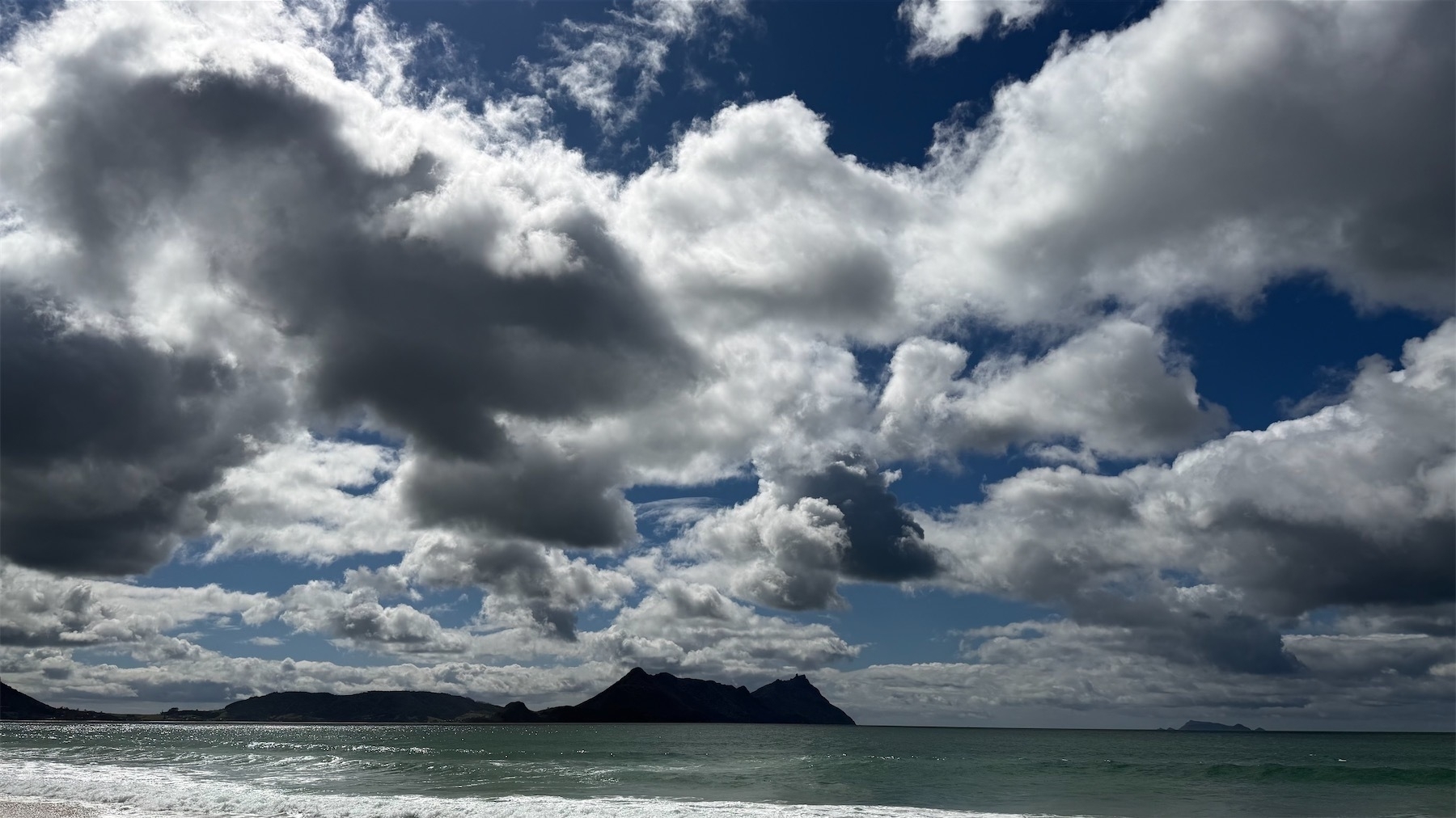 A strip of deep green sea, with hilly backdrop and a dramatically cloudy sky. 