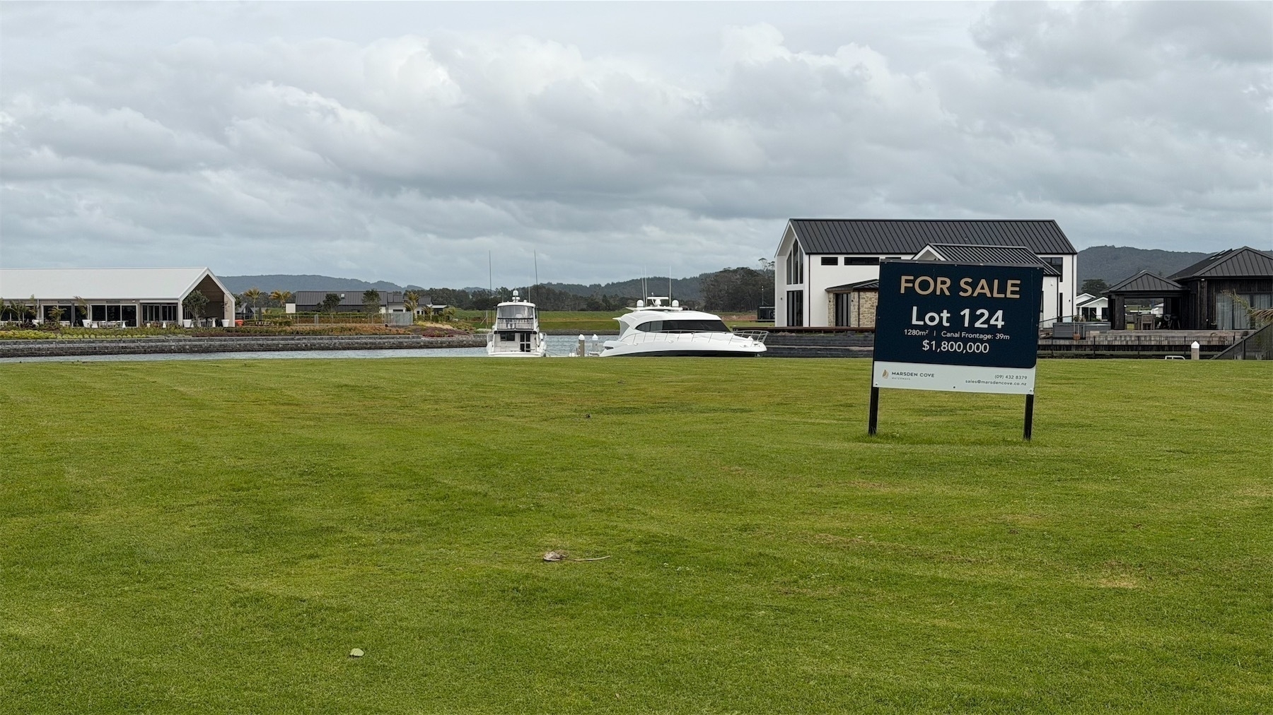 A for sale sign on 1200 square metres of grass with water behind shows a sale price of $1.8 million. In the background are houses and a cafe.