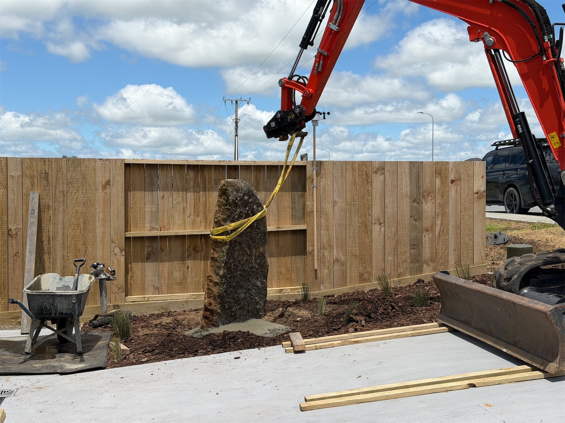 Large rock about 1.2 metres tall with strap attached to a digger and fence behind.