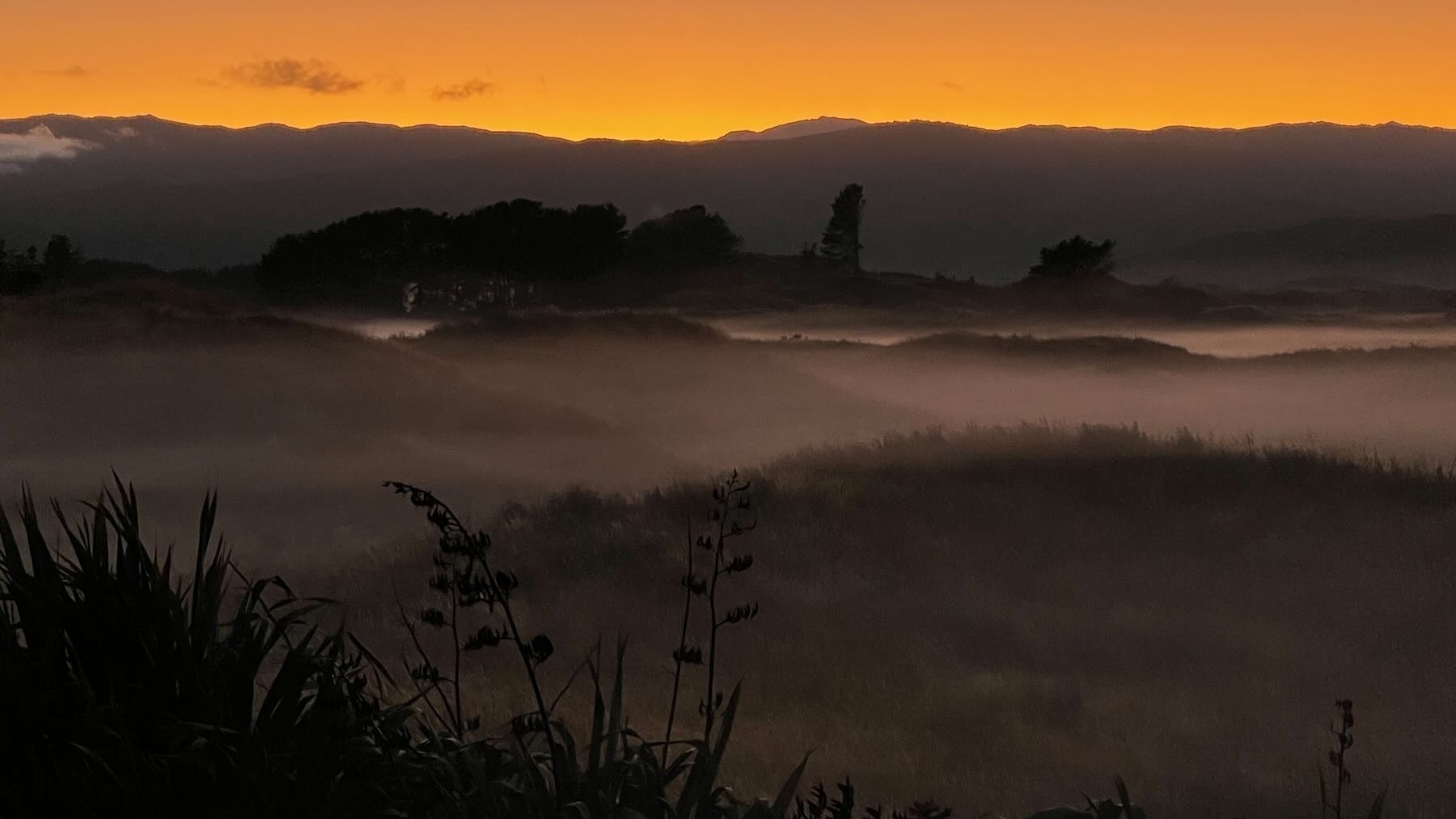 Ground fog across paddocks with dark hills below an orange sky.