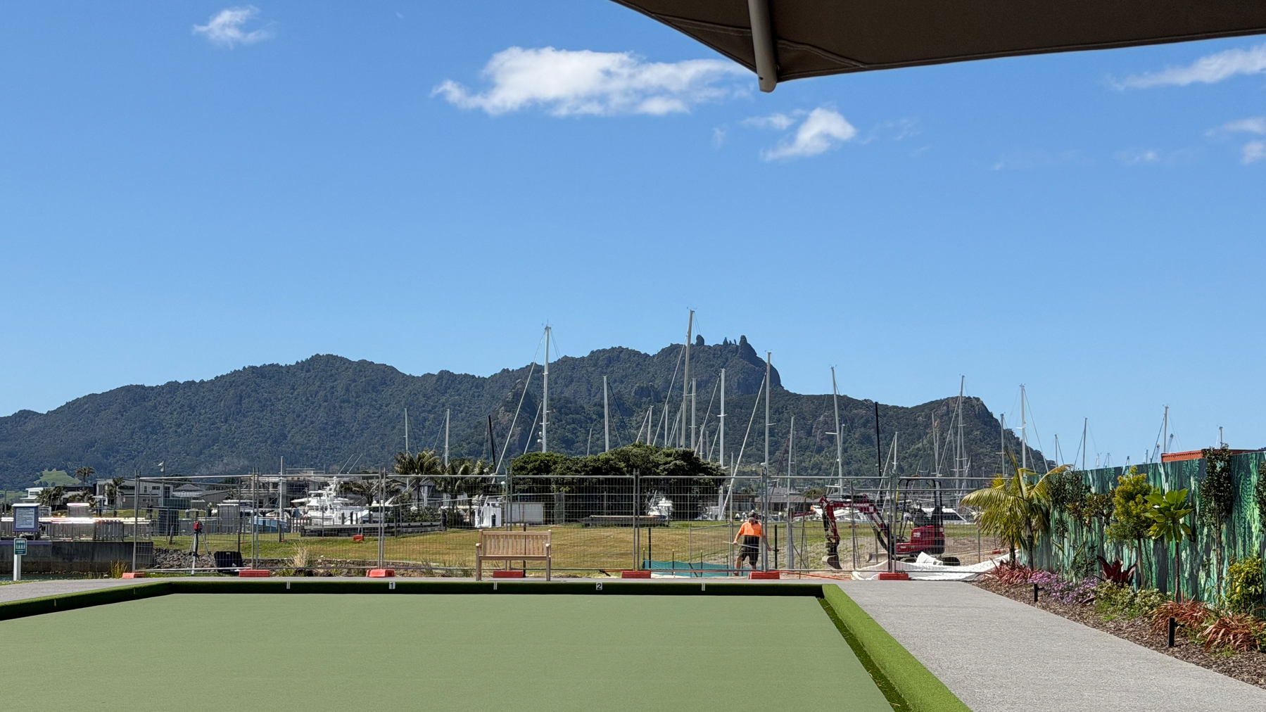 A neatly maintained lawn bowling green is situated in front of a marina with sailboats, set against a backdrop of scenic mountains under a blue sky.