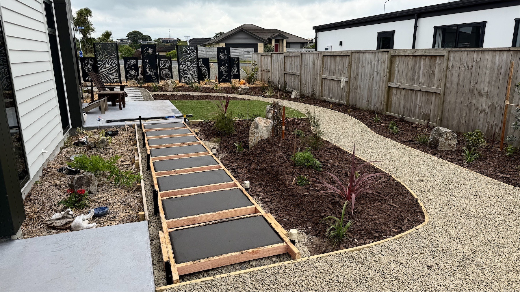 General view of the garden, with a concrete paver path connecting the patios, a winding shingle path, plants on either side.