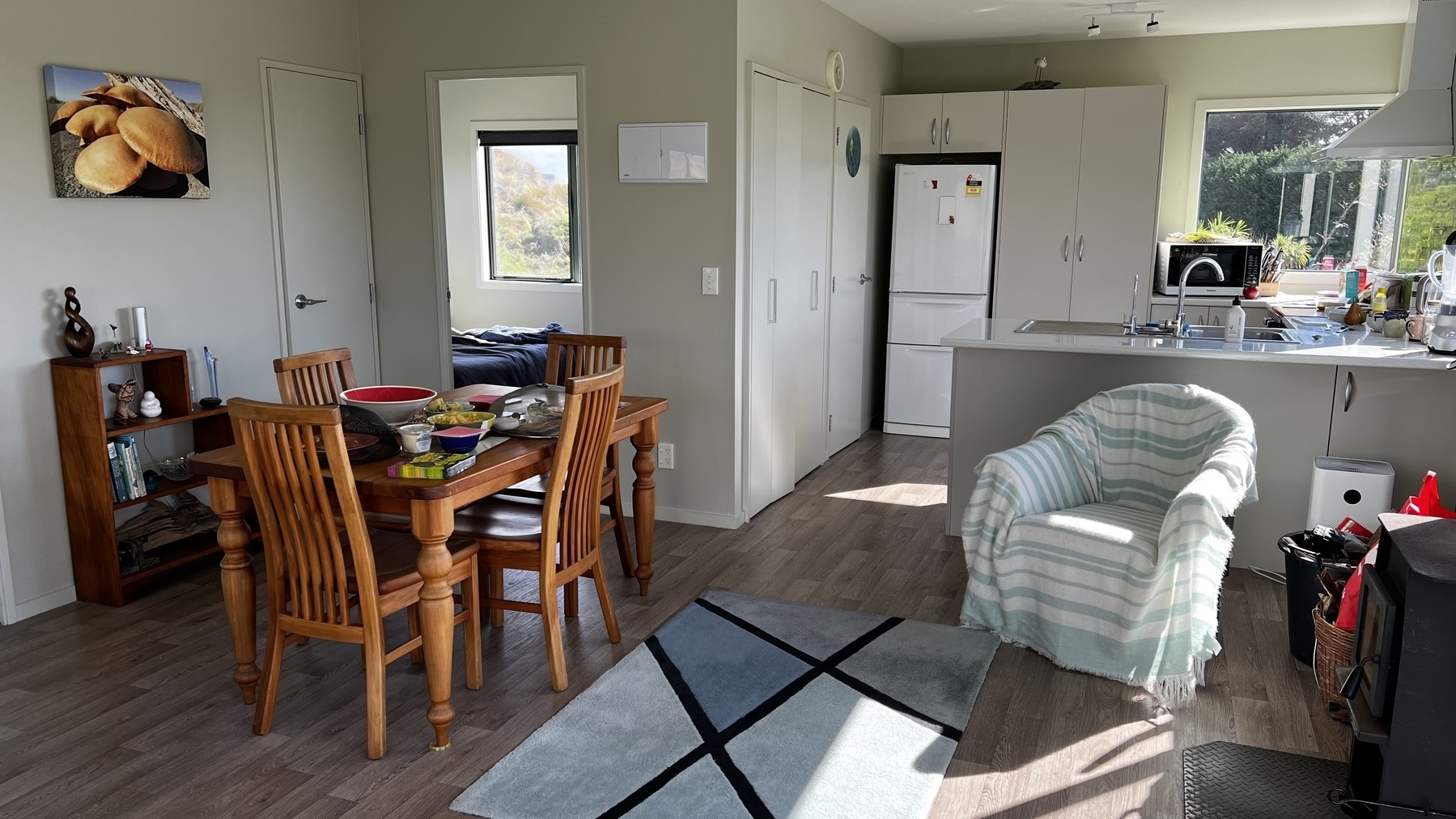 A cozy dining and kitchen area is furnished with a wooden dining set, a cushioned armchair with a striped blanket, and has views of a backyard garden through large windows.