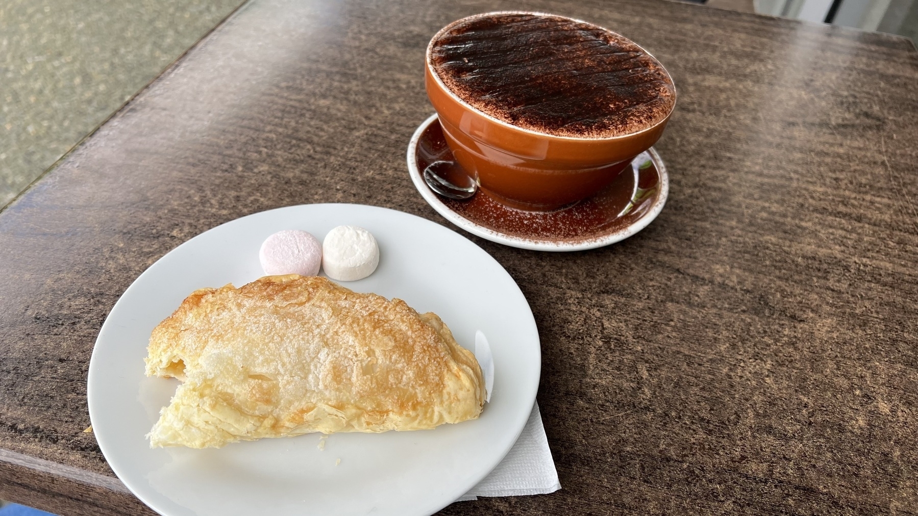 A cup of hot chocolate sits on a wooden table next to a plate with an apple turnover and two marshmallows.