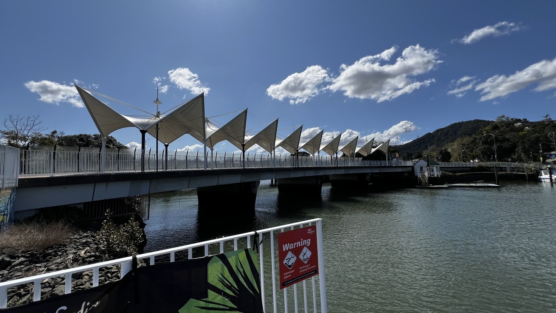 A bridge with triangular canopy structures spans over a body of water under a partly cloudy sky.