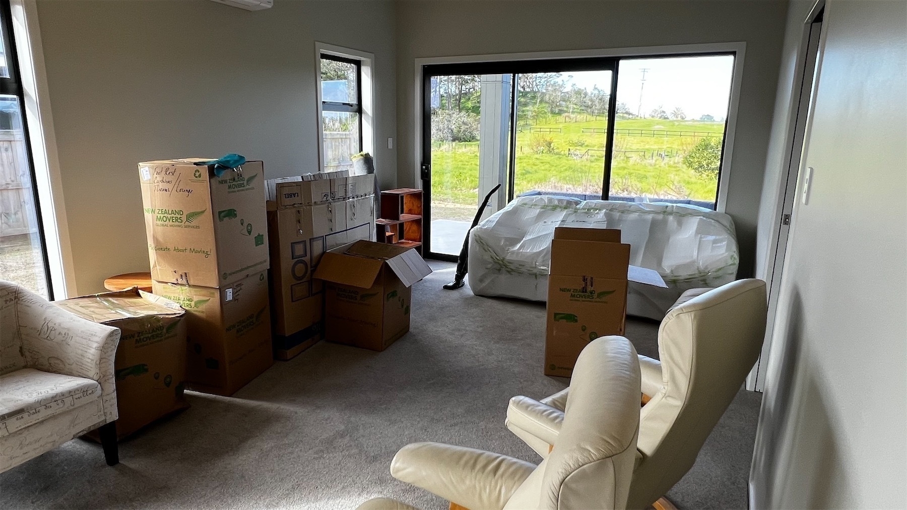 View across a cluttered room, with sofa still wrapped, random furniture scattered around and packing boxes stacked up waiting to be unpacked. 