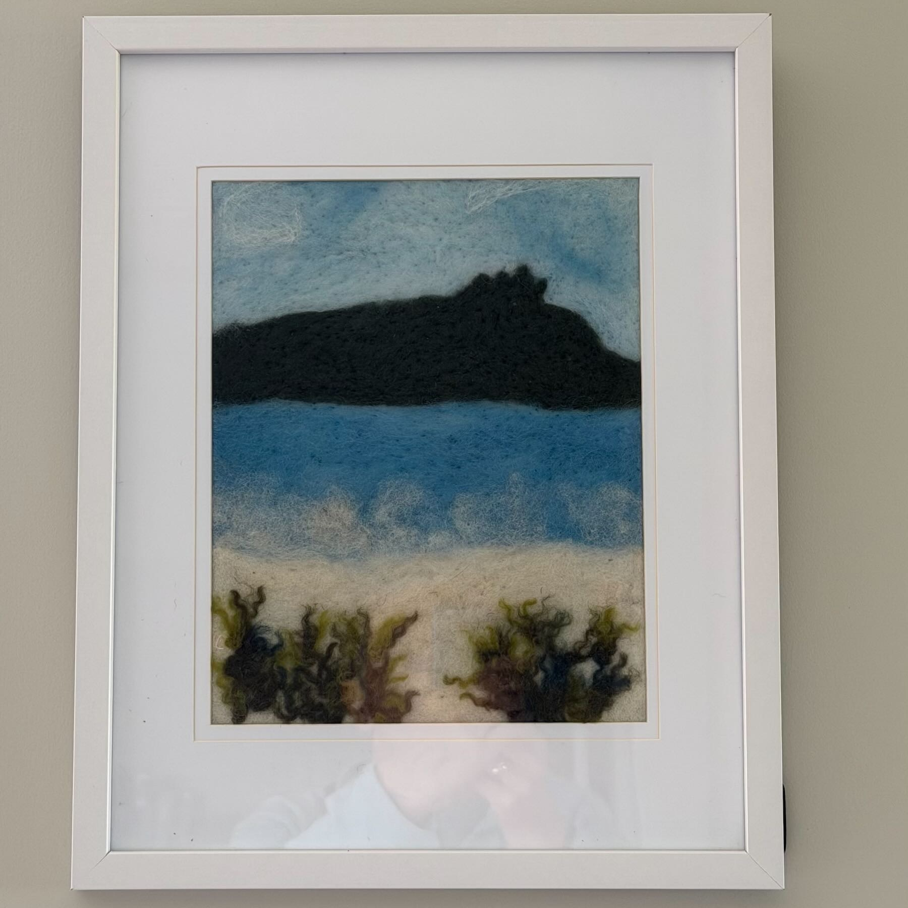 An art work in a white frame depicts a dark hill against pale blue sky, with dark blue water below and white sand with scrubby bushes in front.