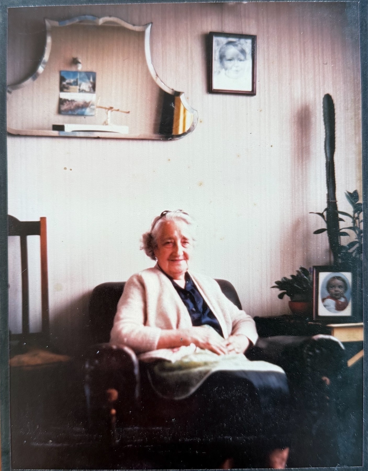 An elderly woman smiling at the camera while seated in an arm chair, with an old-fashioned mirror on the wall behind her head, a tall cactus and some photos on a table nearby. 
