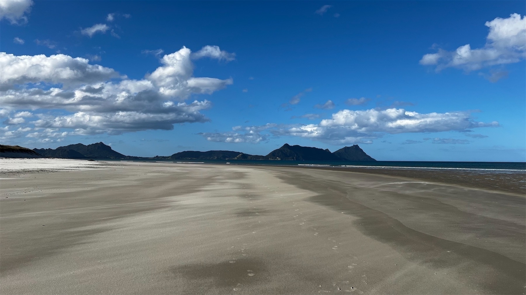 An expanse of sand with dark blue sea on the right and hills not far in the distance. Some puffy white clouds in a blue sky.