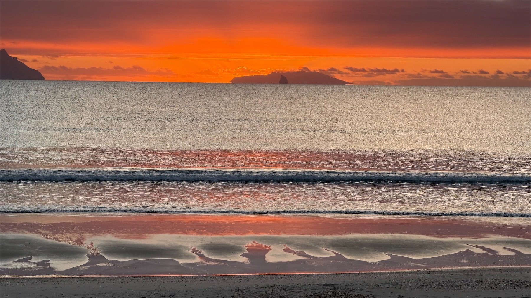 Red sky over silver sea with islands in the background and a tiny wave at the shore.