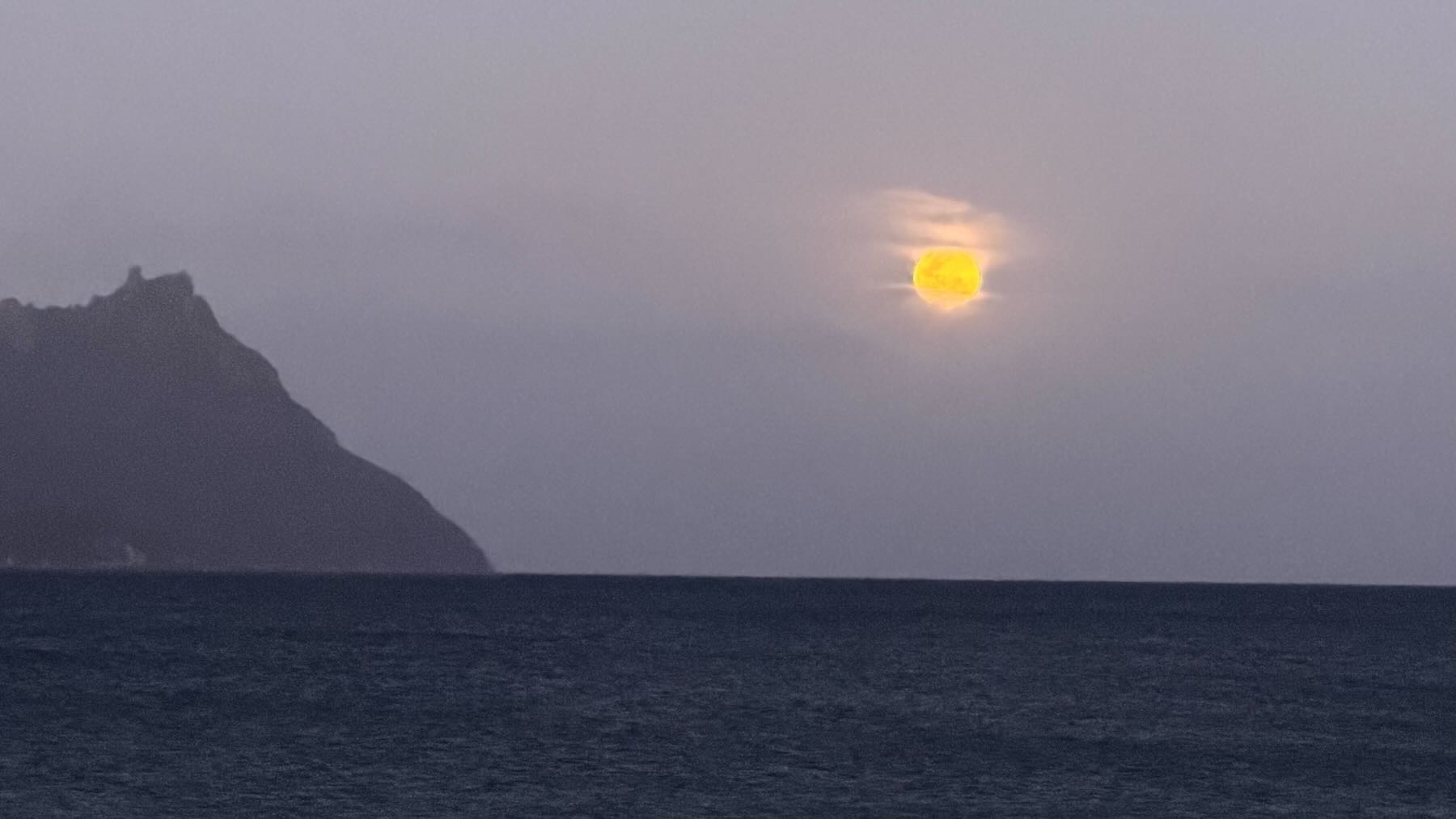 Fiery looking yellow moon with a halo of cloud, above a dark sea and with a mountain nearby. 