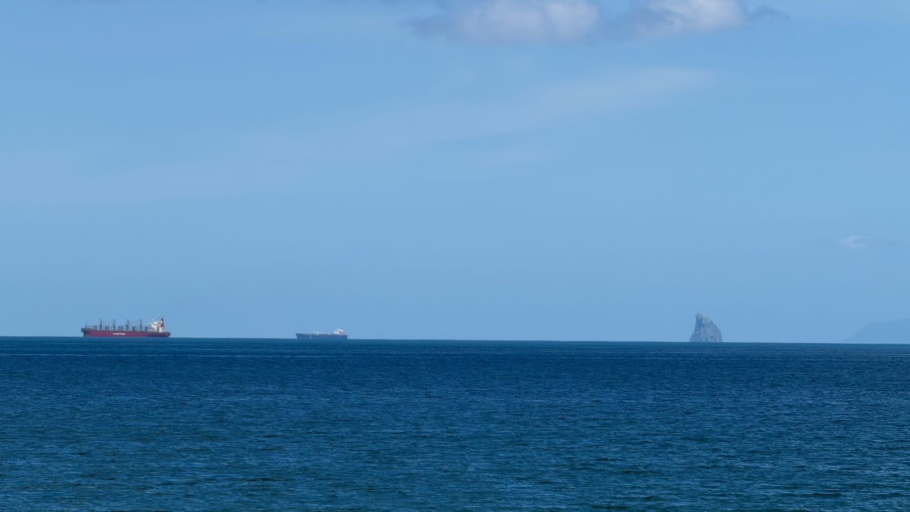 Large ships on the horizon near a large sail shaped rock. 