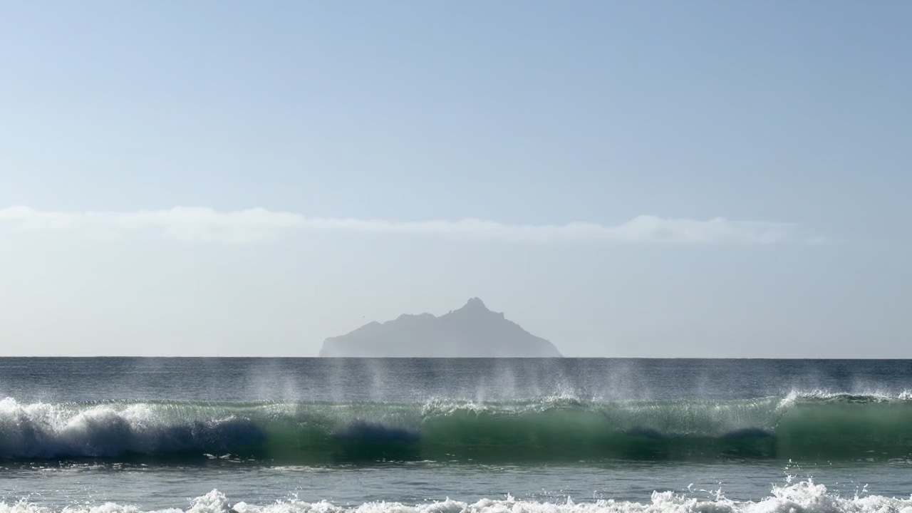 Spray comes off the top of a wave with a distant large island in the background. 
