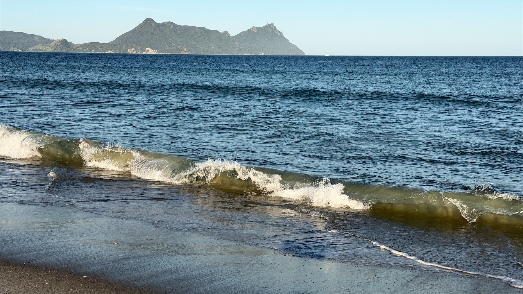 Tiny wave breaks on the shore, with hills and a blue sky in the background.