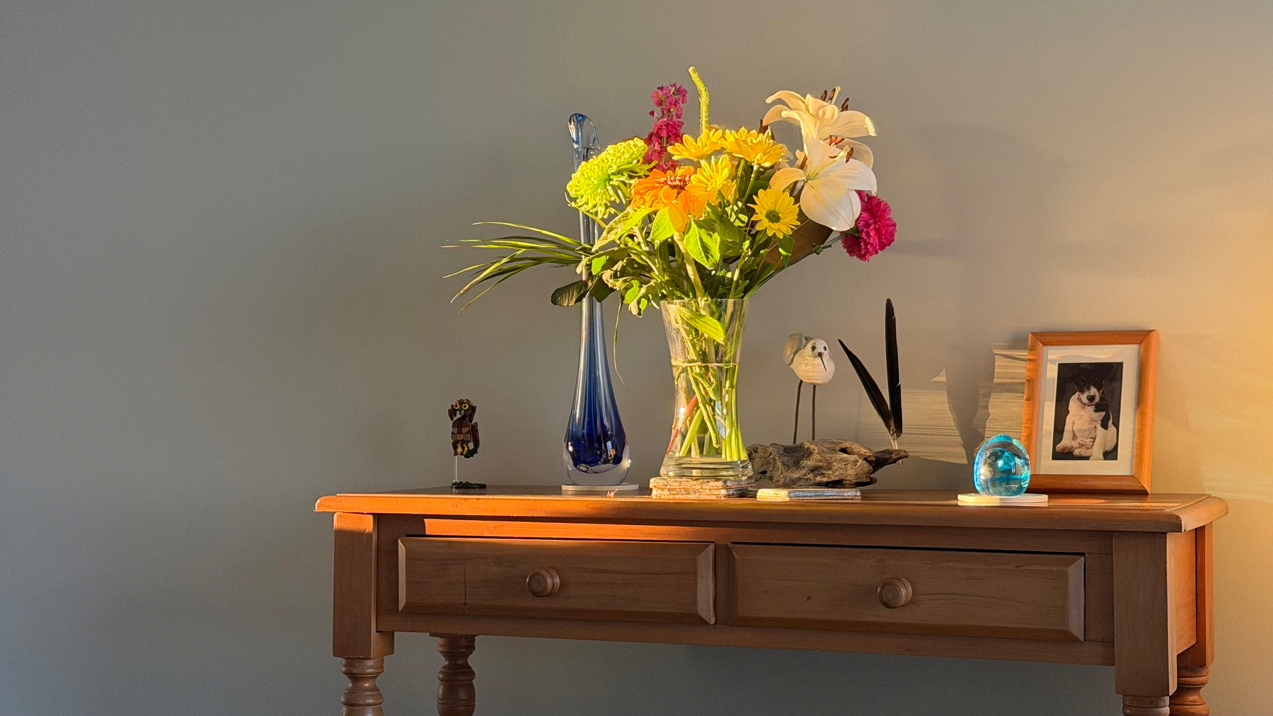 A wooden table holds a colorful bouquet of flowers in a glass vase, a tall blue vase, a framed photo, and several decorative items against a neutral wall.