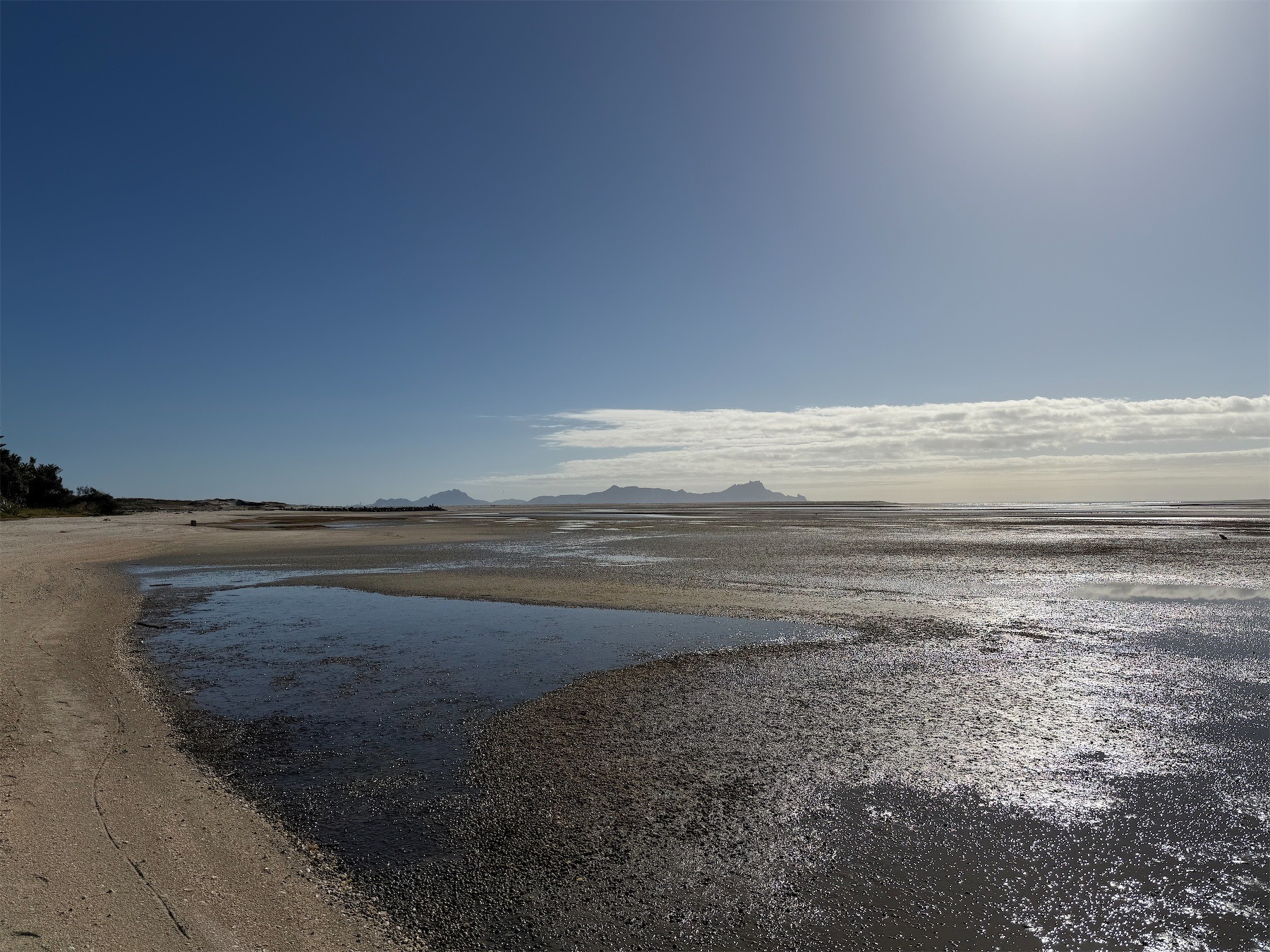 View to the north across wet and dry sand to distant hills. The sun is just visible at top right and the sky is blue. 