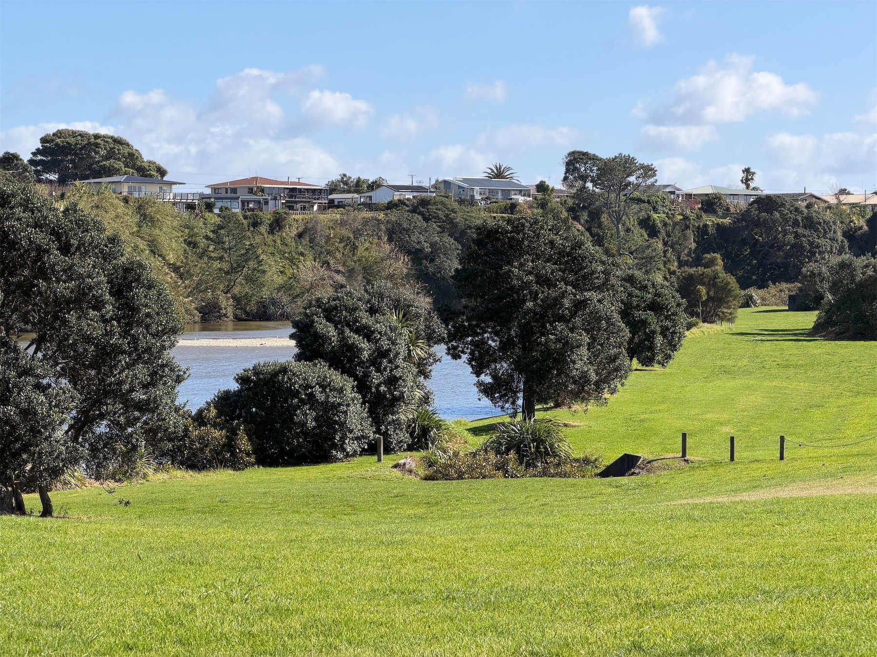 View across grass to a river with trees on the banks and behind a cliff with houses on top. 