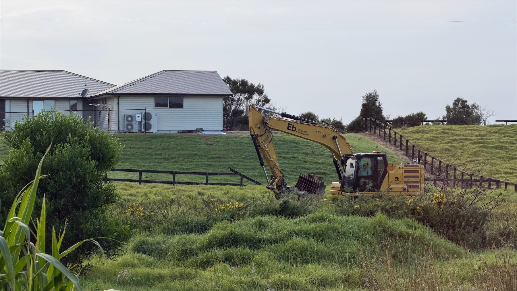 A yellow digger working amongst grass, with low wooden fences and a house in the background.