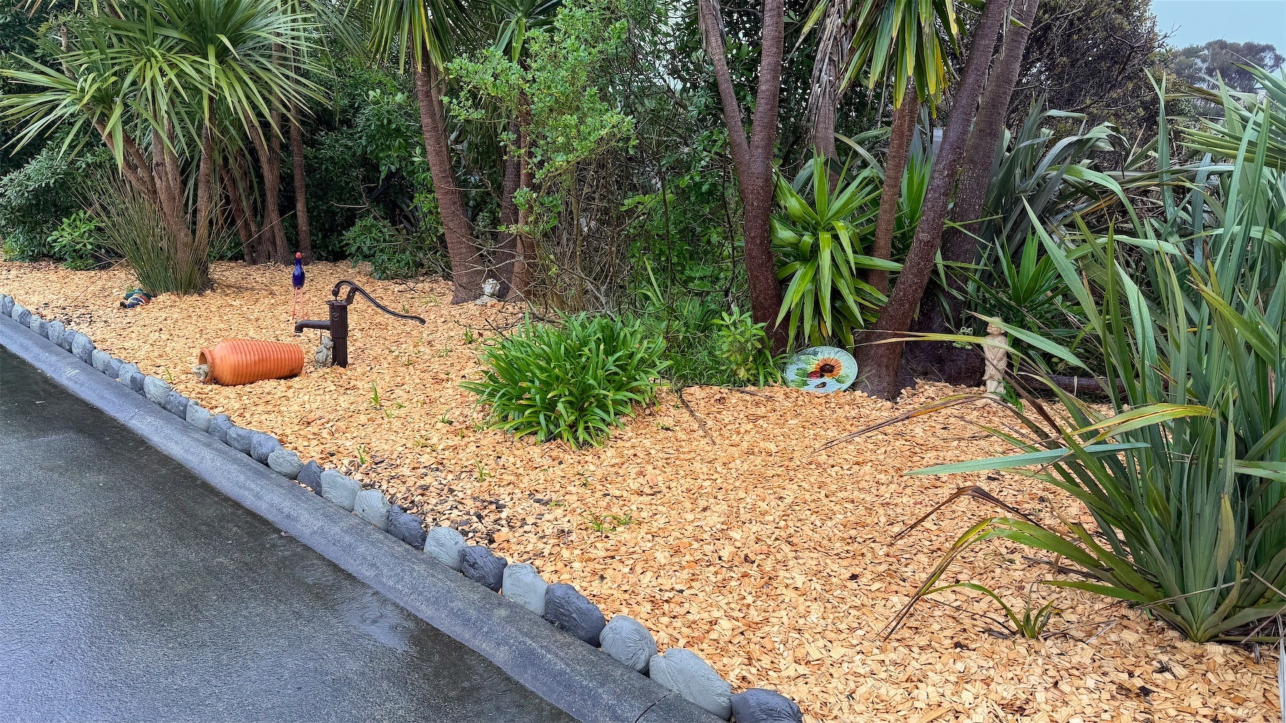 A garden edged with small rocks; mature trees and bushes are at the back; light coloured mulch covers the soil; some small artworks are scattered around.