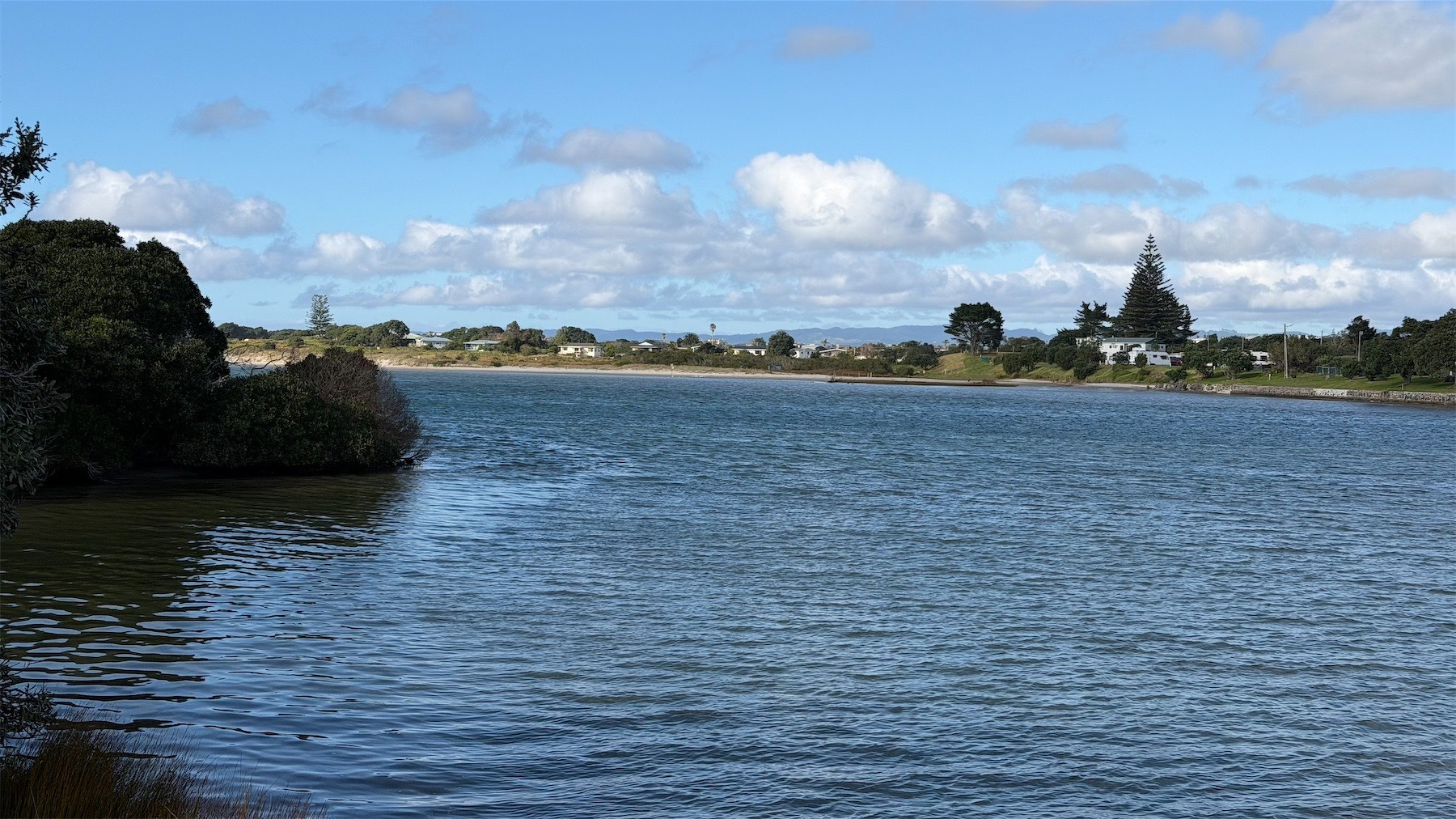 High tide at the mouth of the Ruakākā River, on the far side are trees and small buildings. 