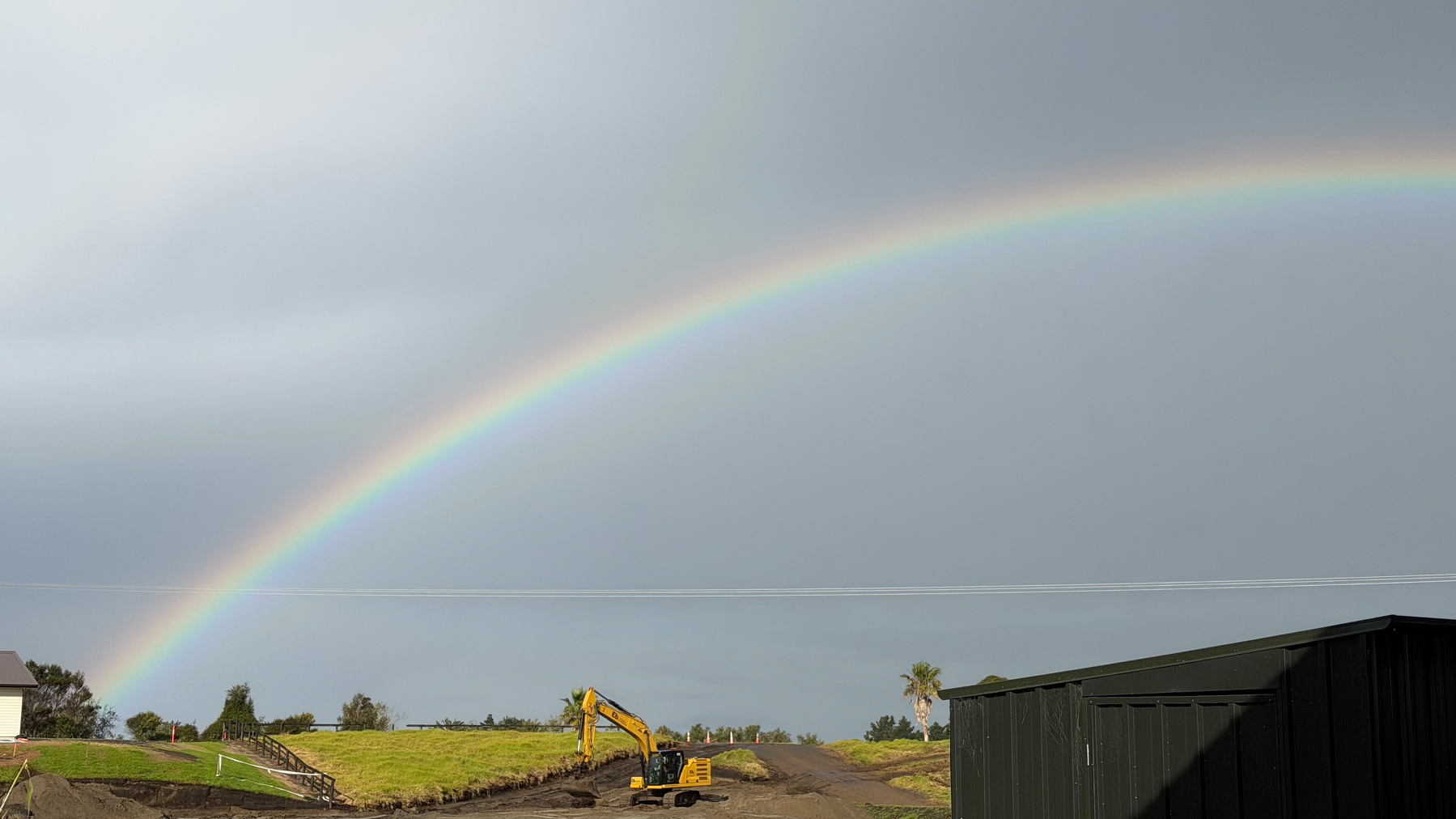 A vibrant rainbow arches over a grassy field with a construction vehicle and a shed under a cloudy sky.
