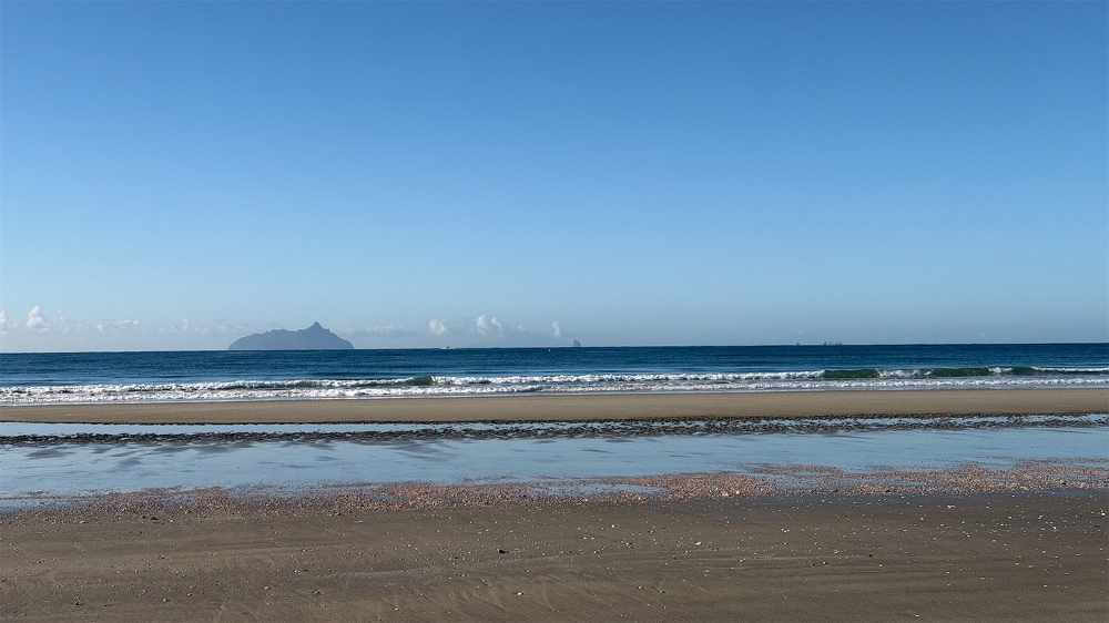 Deep blue calm sea behind an expanse of sand, blue sky and islands behind. 